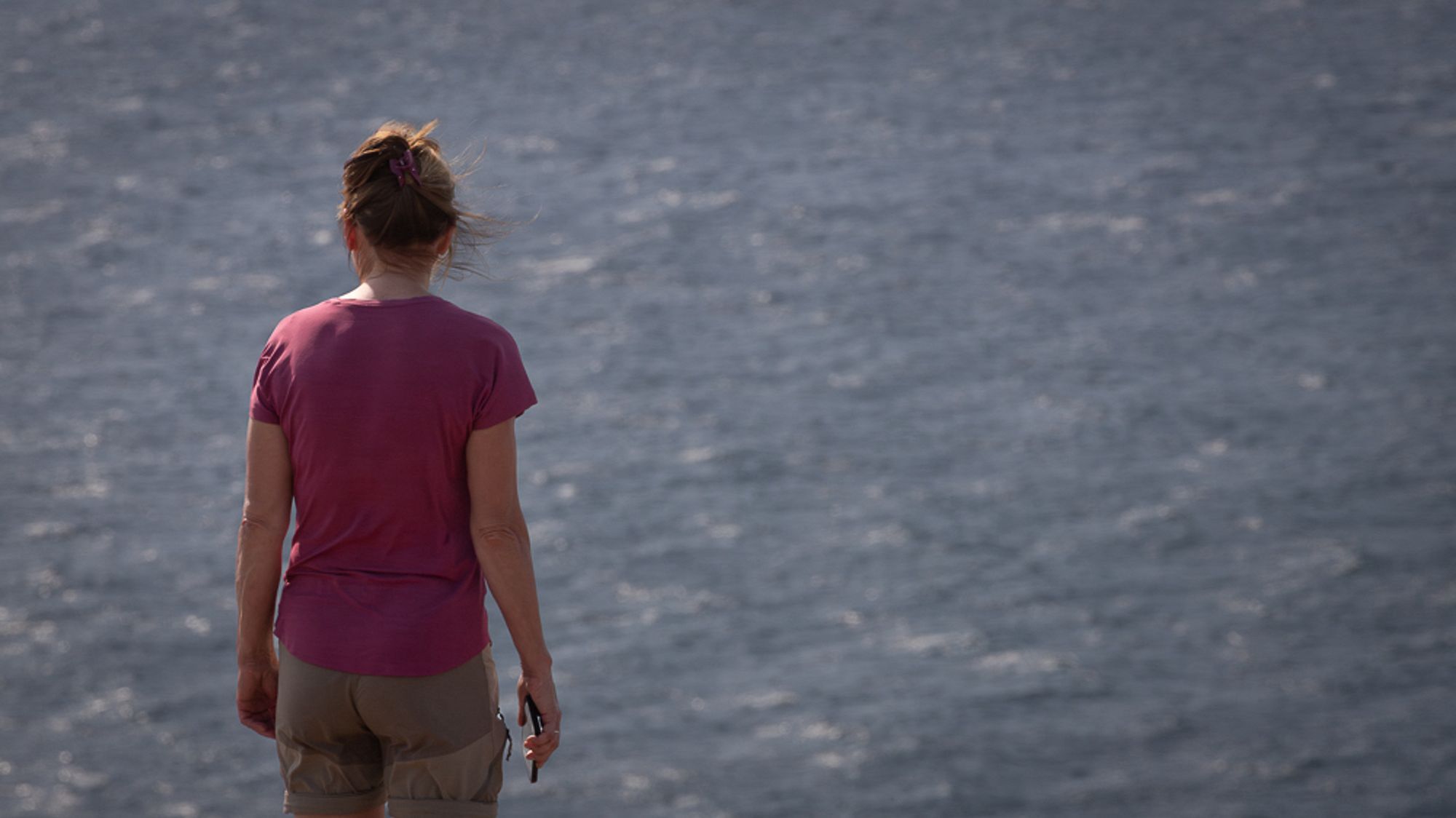 A person stands with their back to the camera, overlooking a body of water. They are wearing a short-sleeved, pink shirt and light-colored shorts. The scene captures a serene atmosphere with a soft breeze.