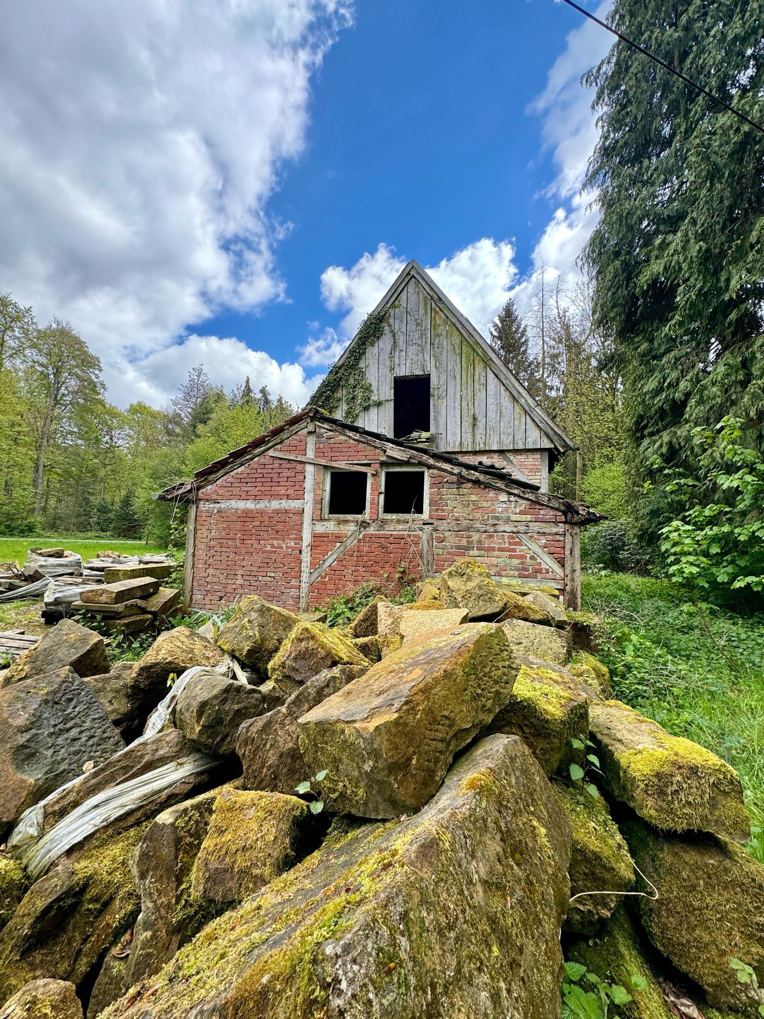 An old dilapidated barn with a moss-covered stone pile in the foreground and dense trees in the background under a partly cloudy sky.