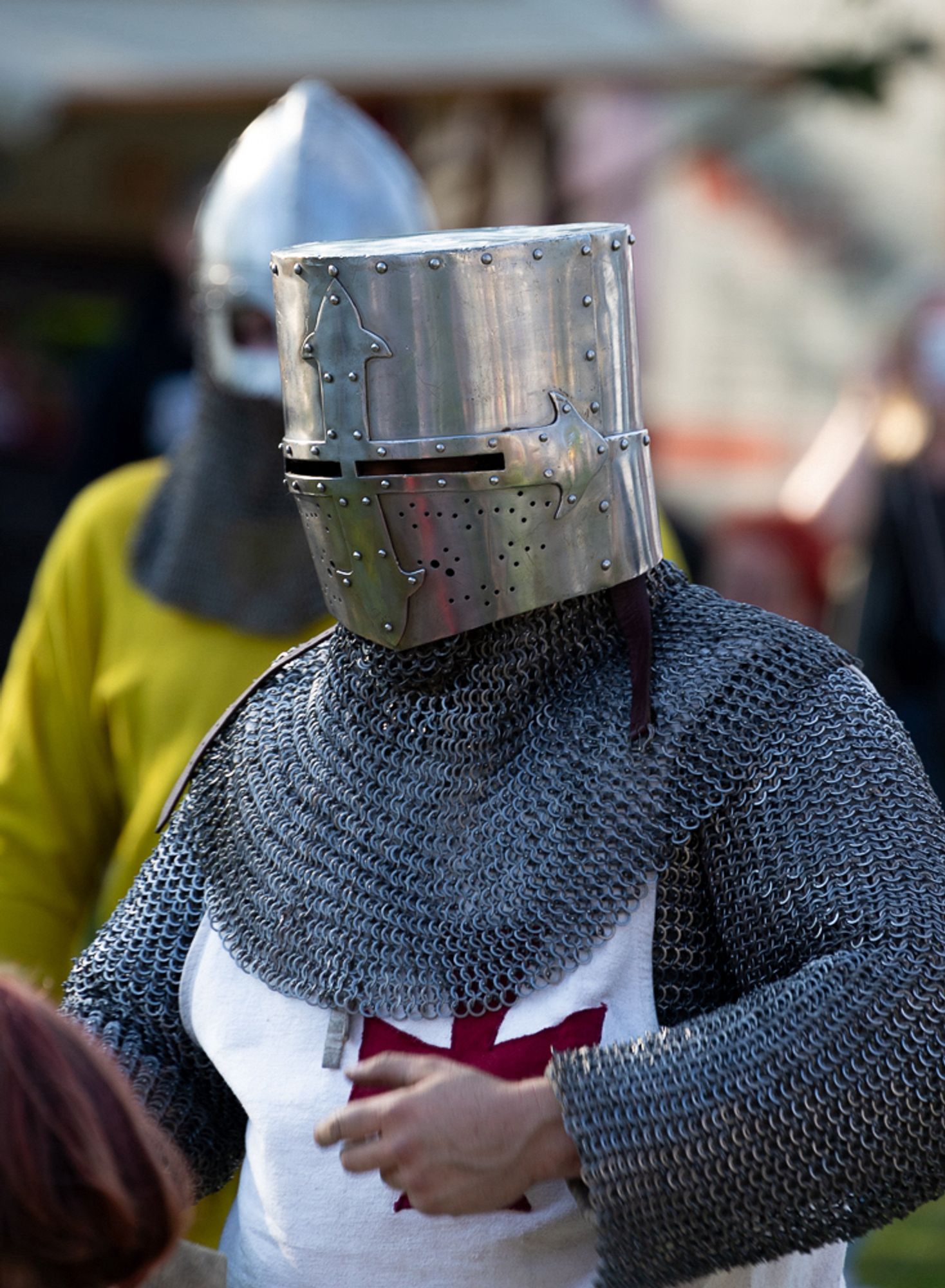 A person wearing medieval knight armor, including a chainmail suit and a metallic helmet with a cross symbol. The person is shown waist upwards.