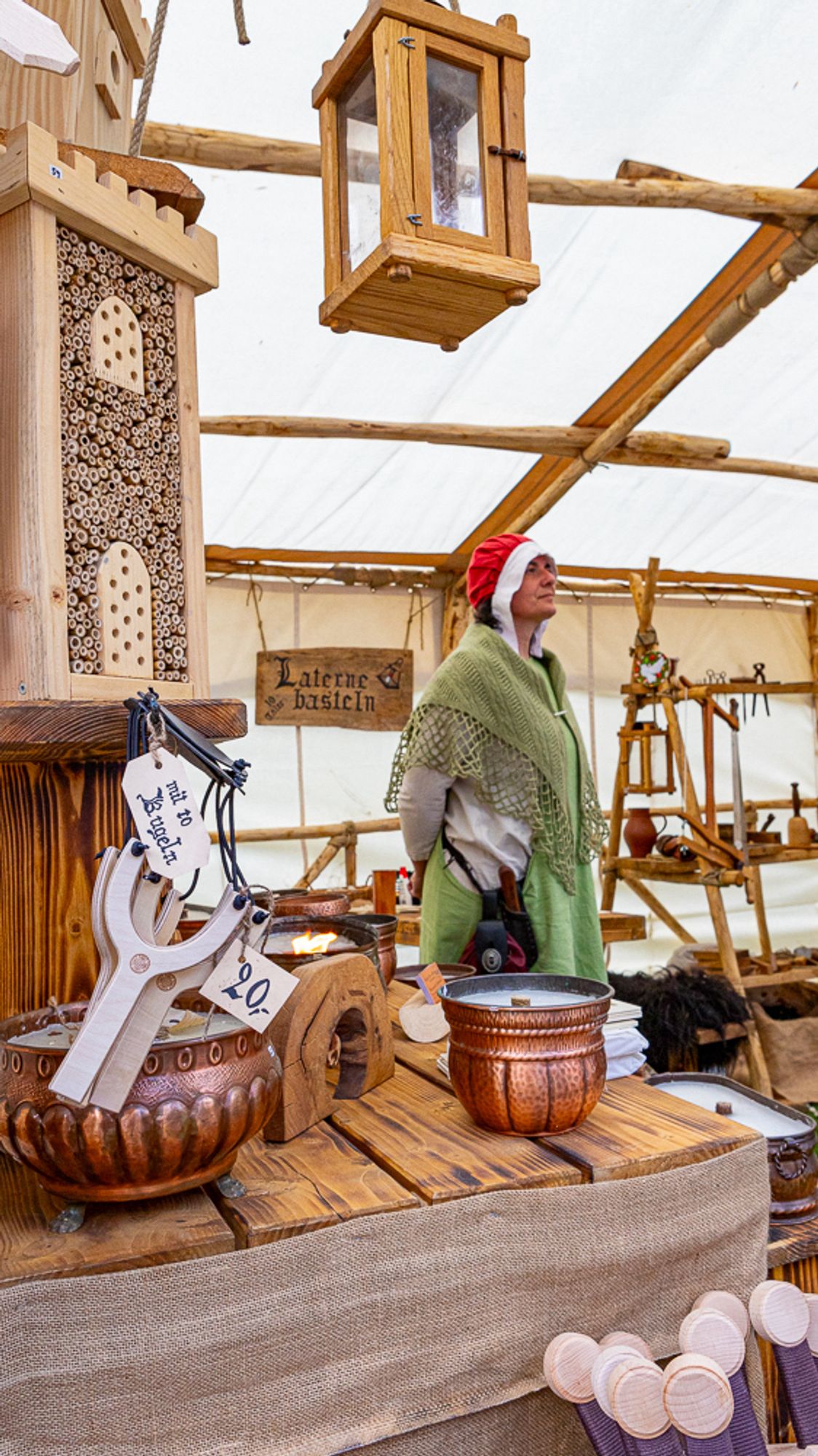 A person in historical attire stands in a wooden craft stall, displaying handmade items such as wooden slingshots, lanterns, and bowls.