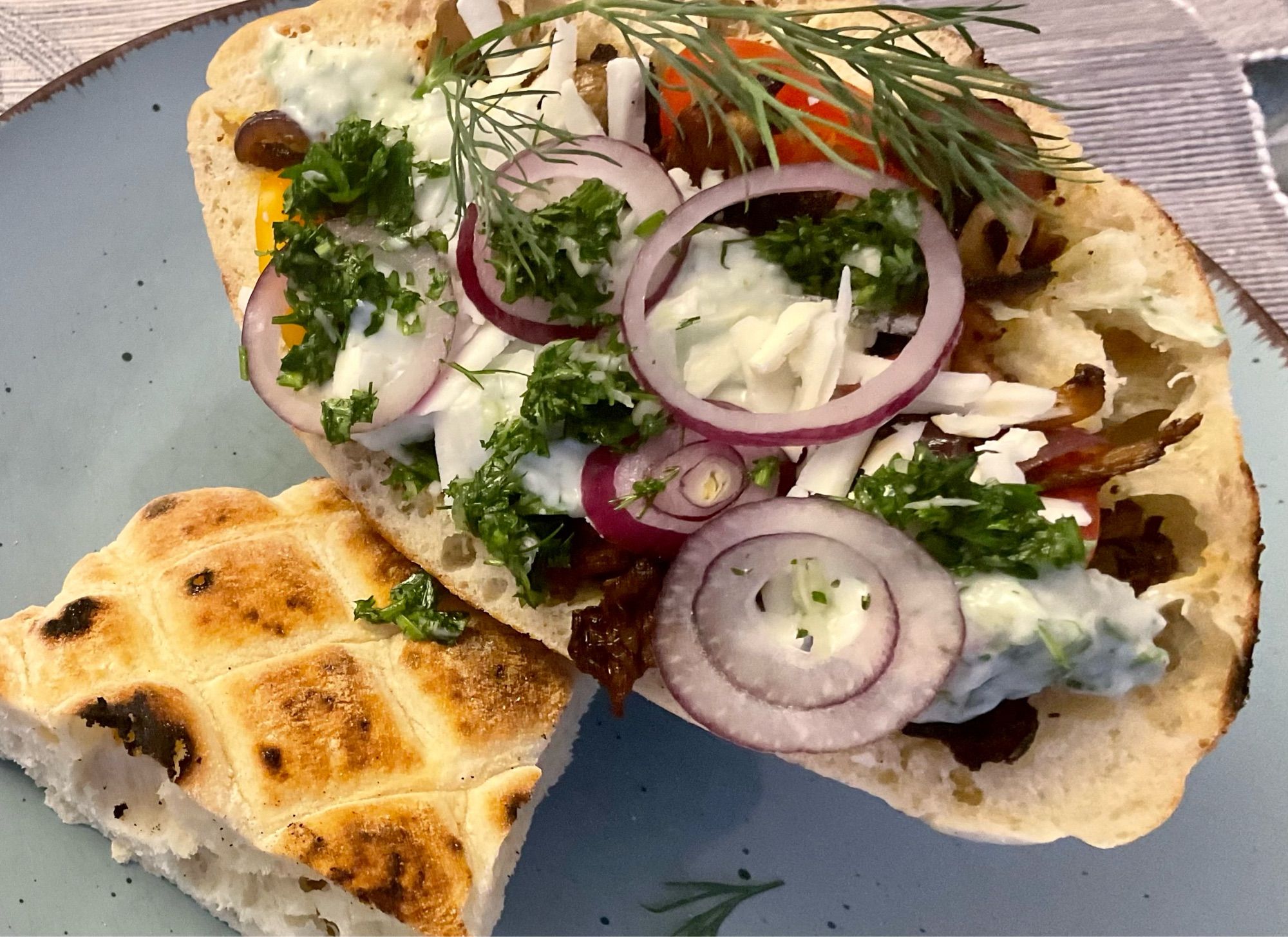 Auf einem grauen Teller befindet sich Pitatasche gefüllt mit Pilz Gyros,veganem Tzatziki,Tomaten,Gurke und veganem  geriebenem Feta.Obendrauf würde das Gericht mit Zwiebelringen und Dill dekoriert