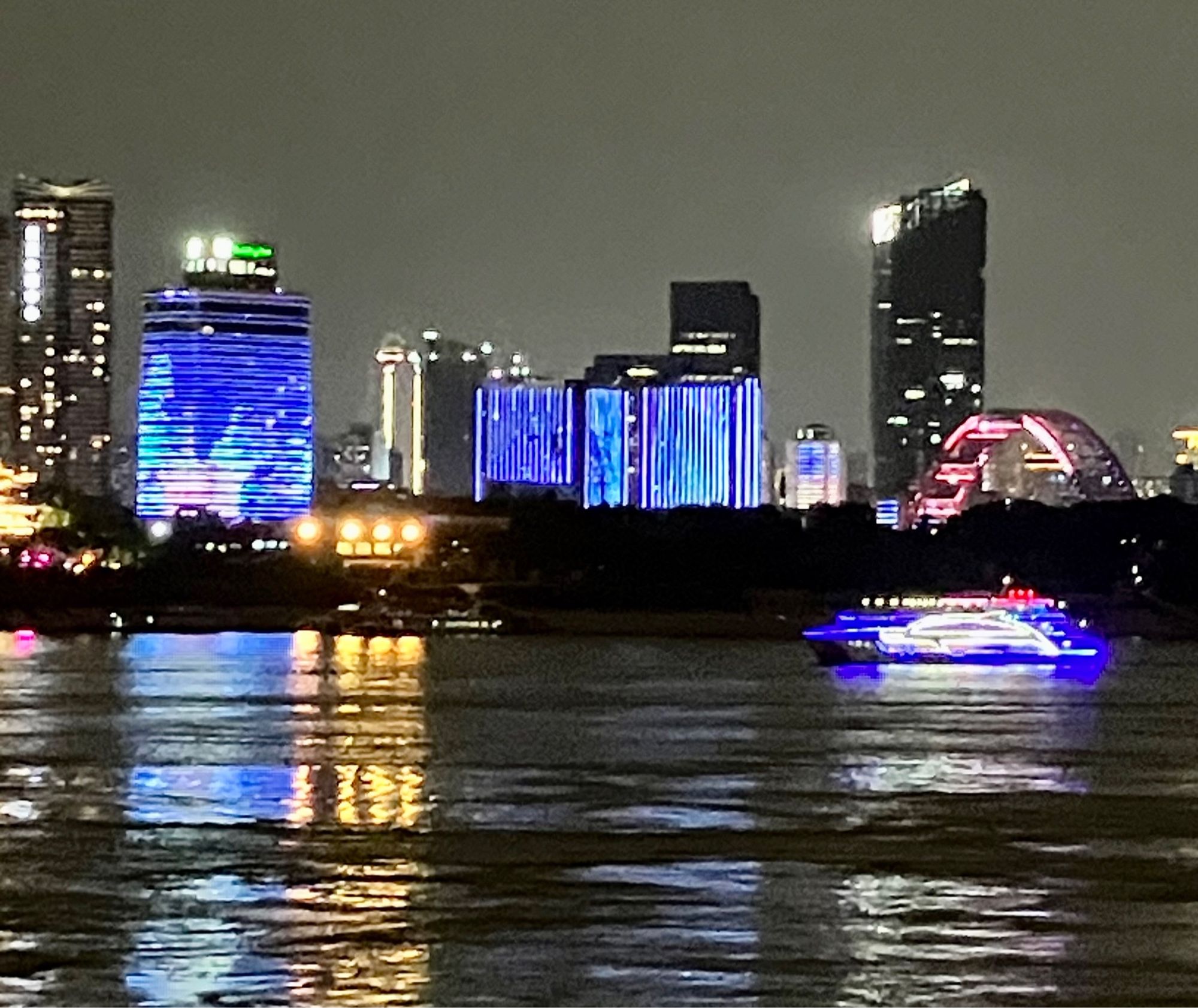 Yangtze River at night with large skyscrapers lit up blue in the background. A tour boat covered in lights shaped like a dolphin is passing by