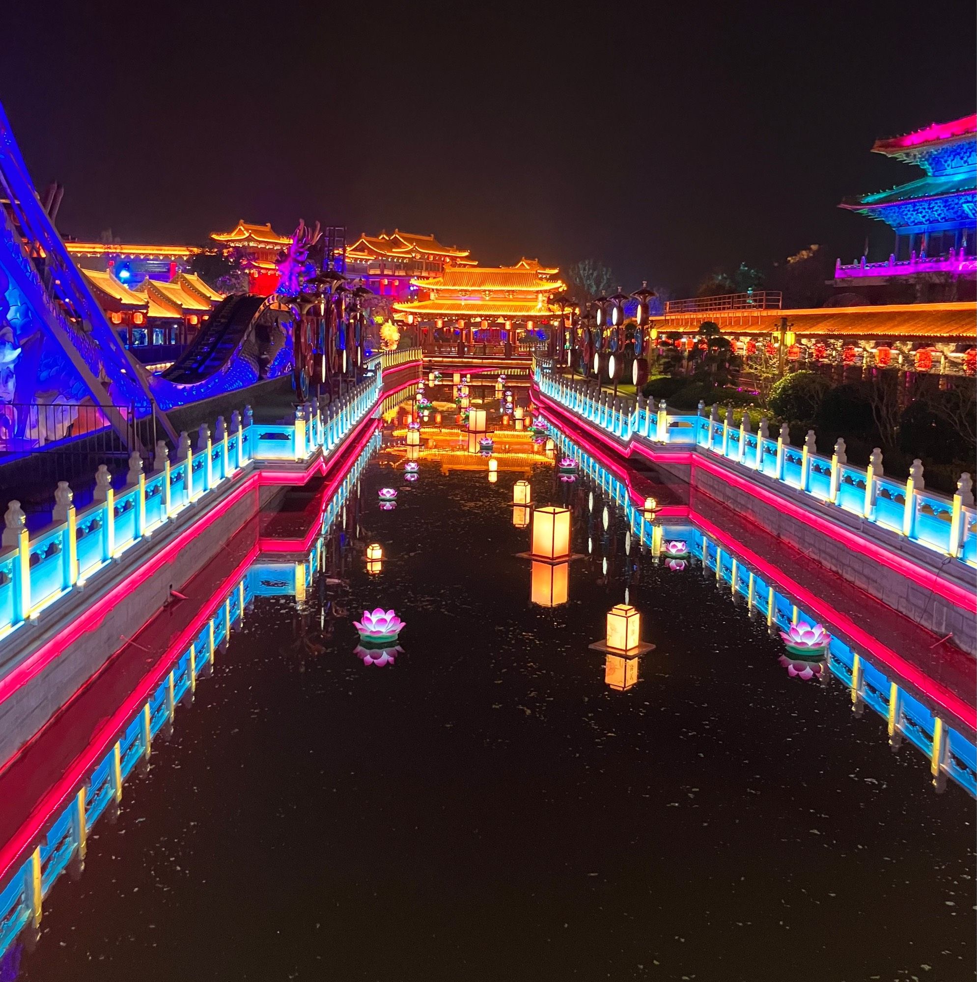 Lights lining a canal with lit lanterns and a Song era style building lit up in the background.