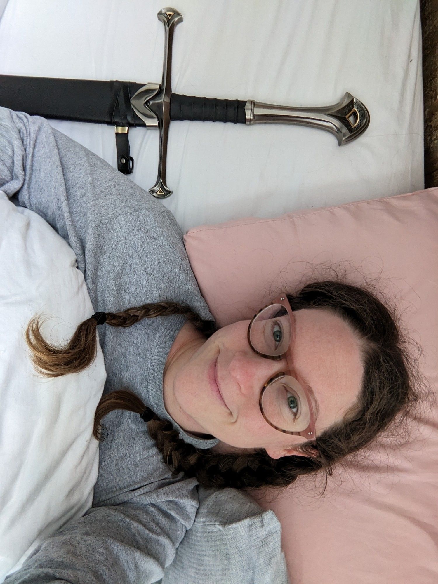 A photo of me (a white woman wearing glasses with my brown hair in two braids) lying in bed in the morning beside my sword, Andúril.