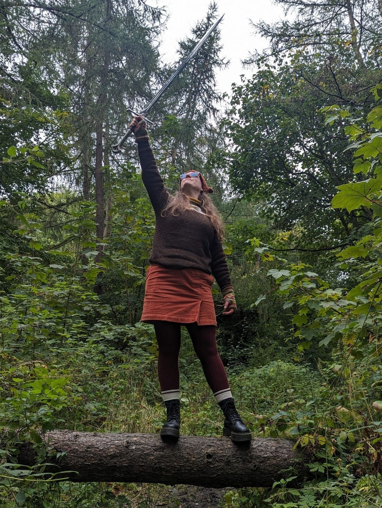 I'm standing on a log in a forest wearing an autumnal outfit of orange, brown, and burgundy, and holding up Andúril, Flame of the West, a massive sword. I look cool.