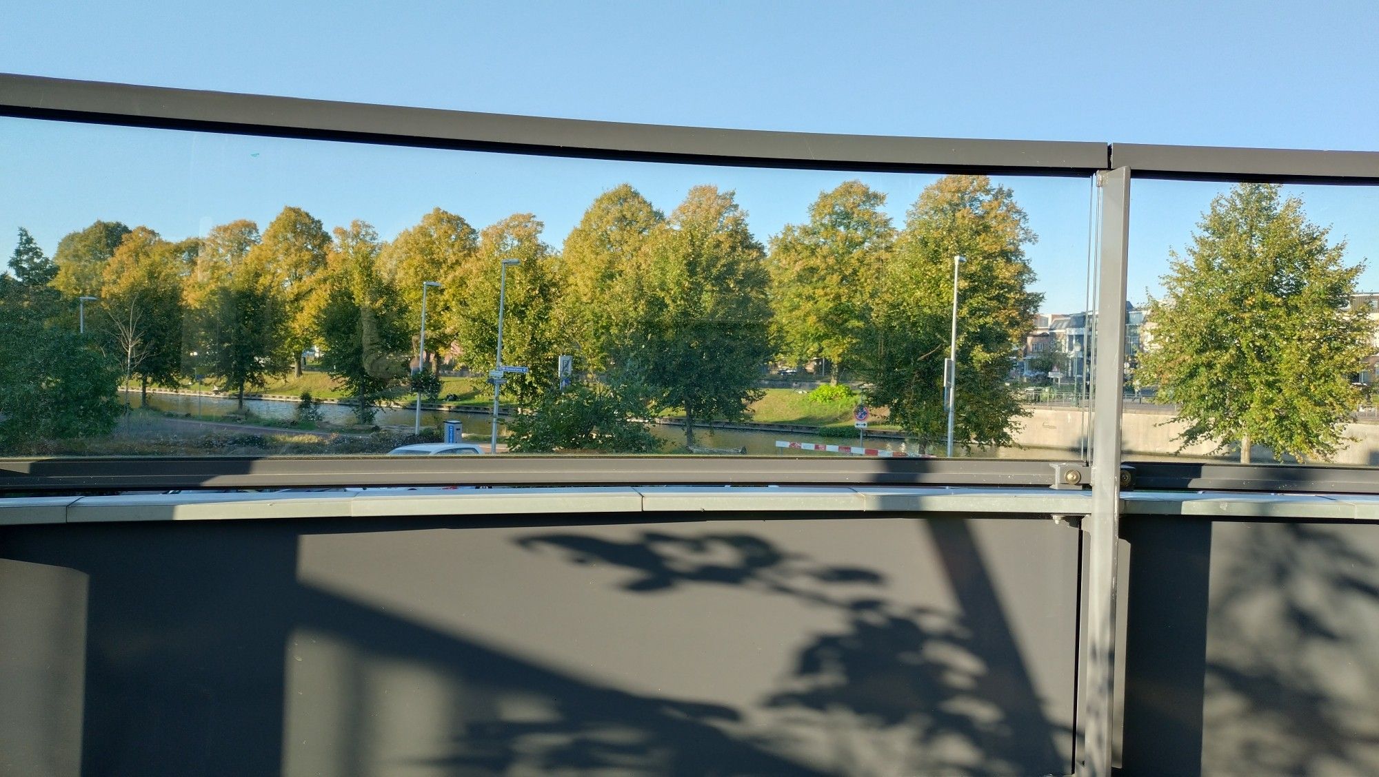 Balkonhek onderkant staal met schaduw plant en bovenkant glas waarachter bomen in beginnende herfstkleuren.