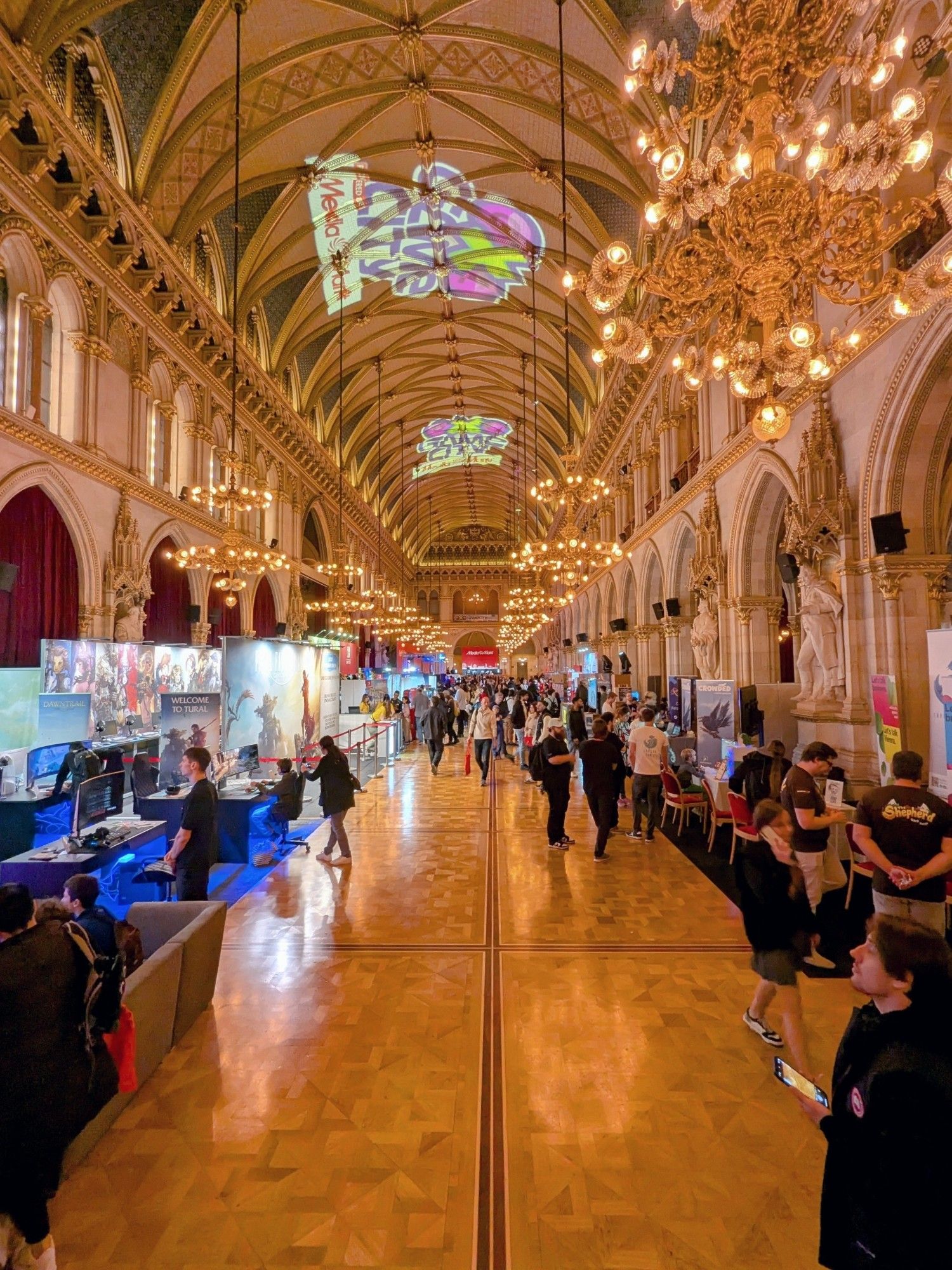 Ein großer Saal im Wiener Rathaus als Gaming Hall umfunktioniert. Man sieht den prunkvollen Raum mit vielen Kronleuchter bevölkert von vielen Personen vor Videospiel-Anspielstationen und Informations-Ständen.