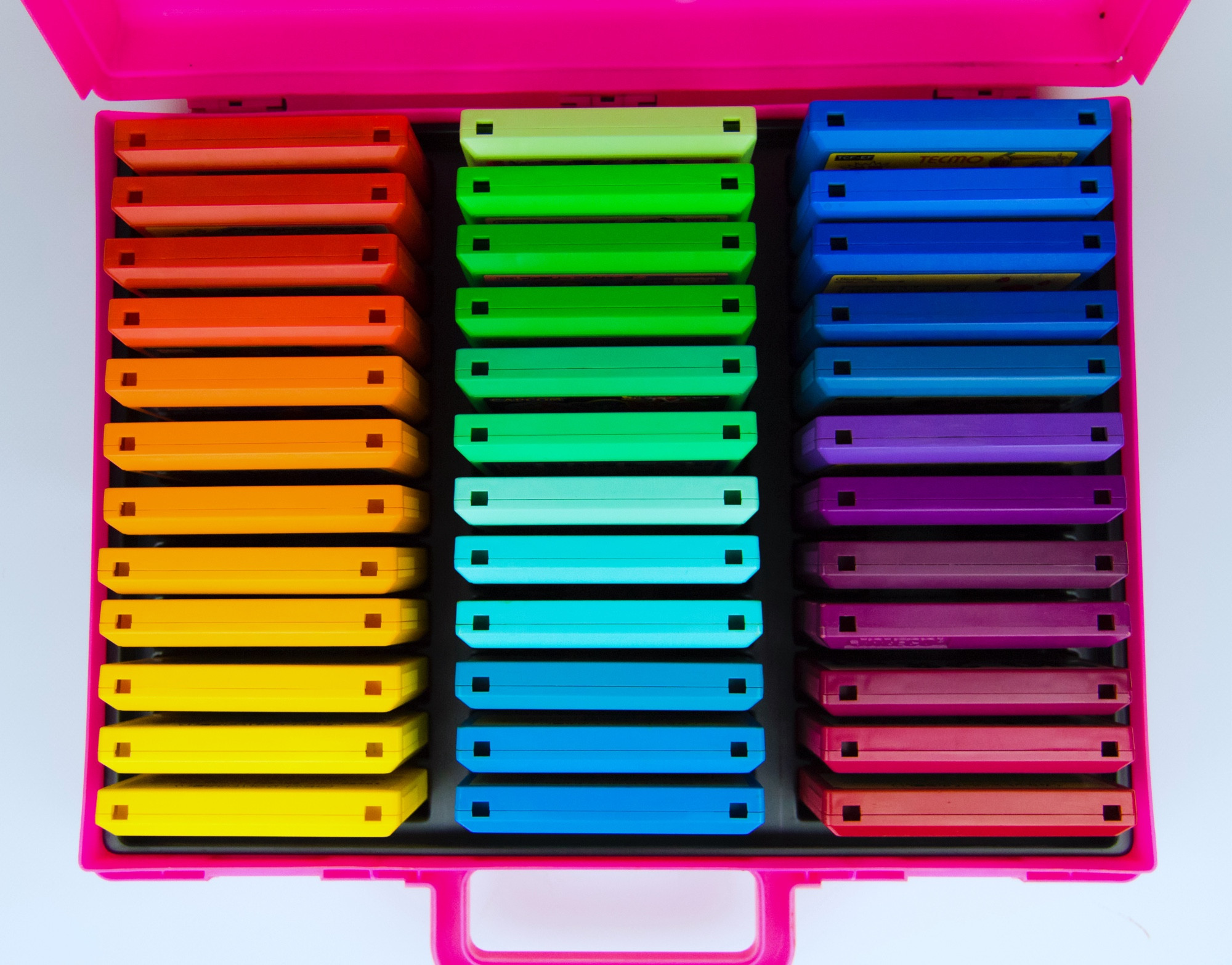 Multicolored Famicom cartridges arranged in a rainbow pattern inside a pink audio cassette storage case