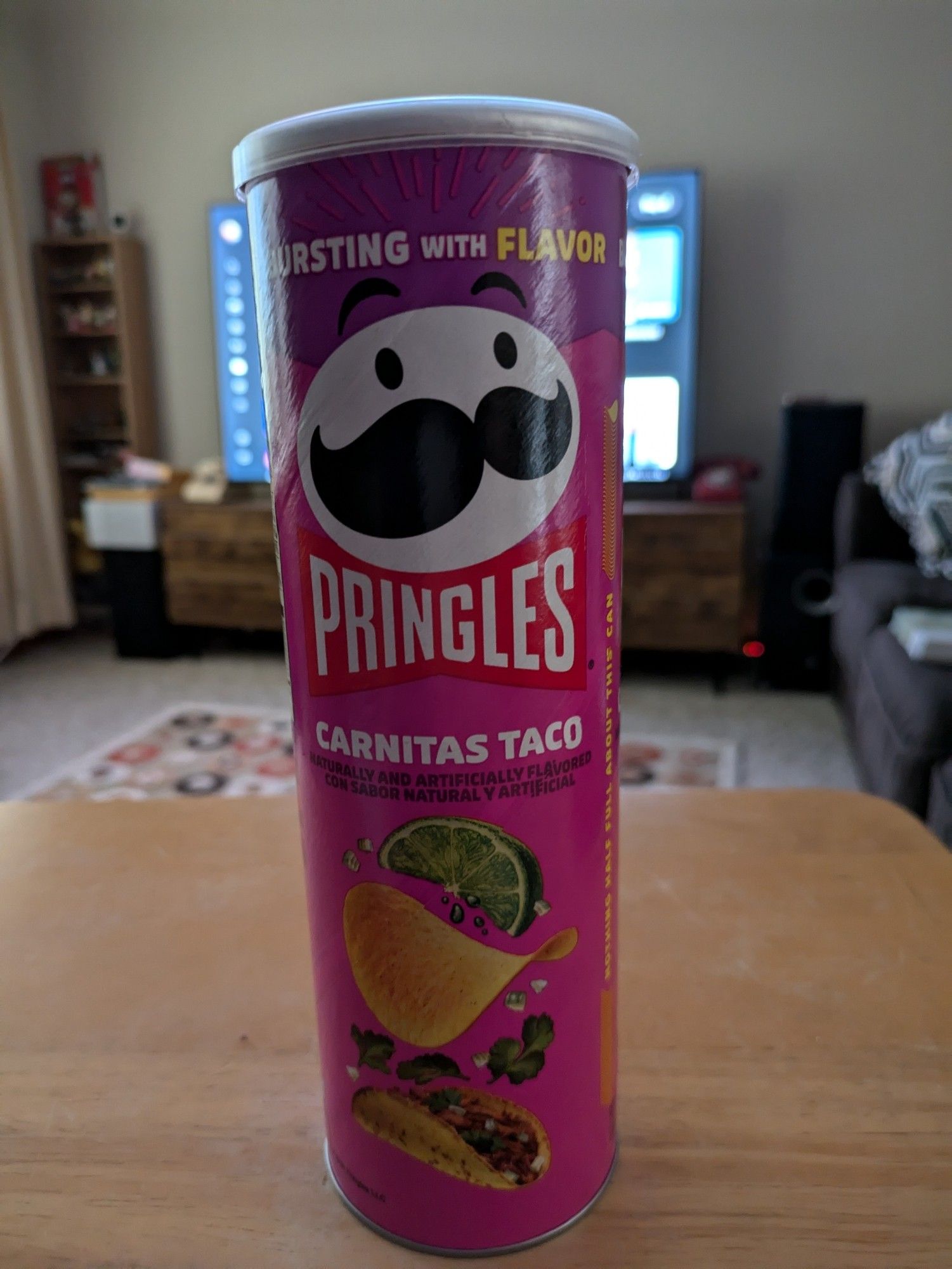 A can of Pringles carnitas taco flavor chips sitting on a tray table. In the background is my living room with a TV on, but obscured by the Pringles can.