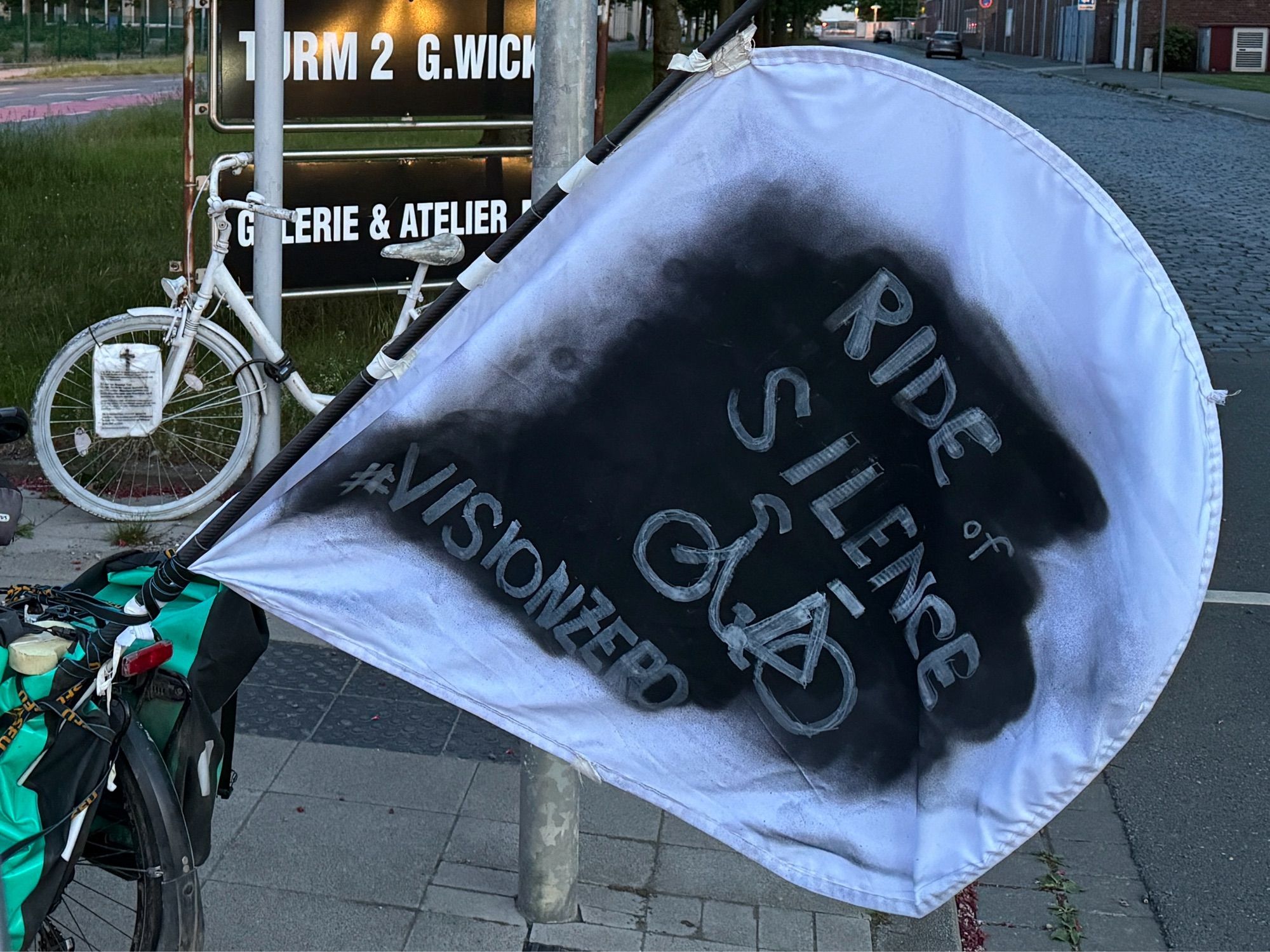 Foto. Im Hintergrund ein weißes Fahrrad. Im Vordergrund eine weiße handgemachte Fahne. Auf einer eingeschwärzten Fläche steht: „Ride of Silence. #visionzero“