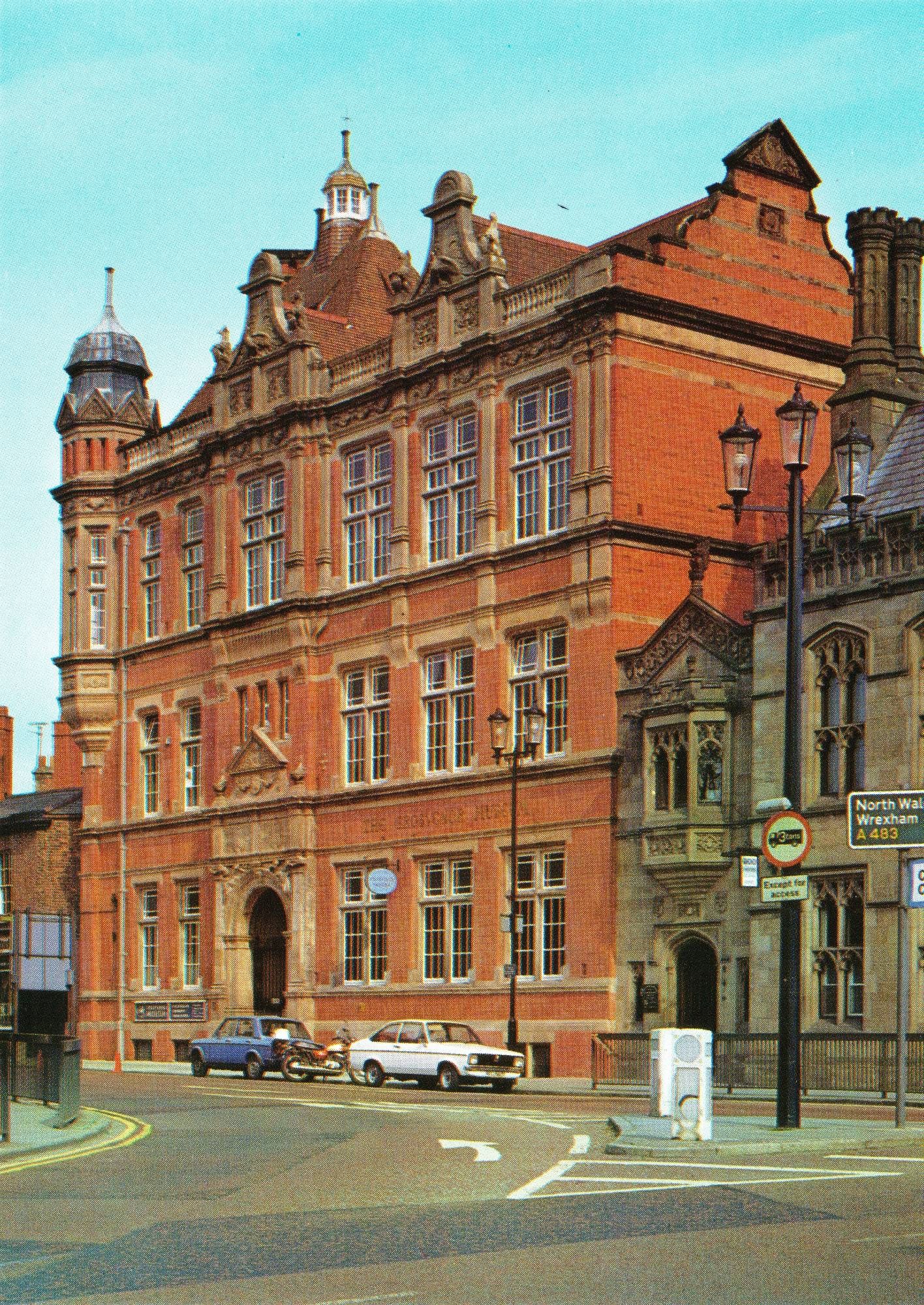 A colour postcard with a photograph of the ornate four storey red brick museum in Chester. There is a lavish entrance on the ground floor, a turret with a cuppola and a lantern tower!