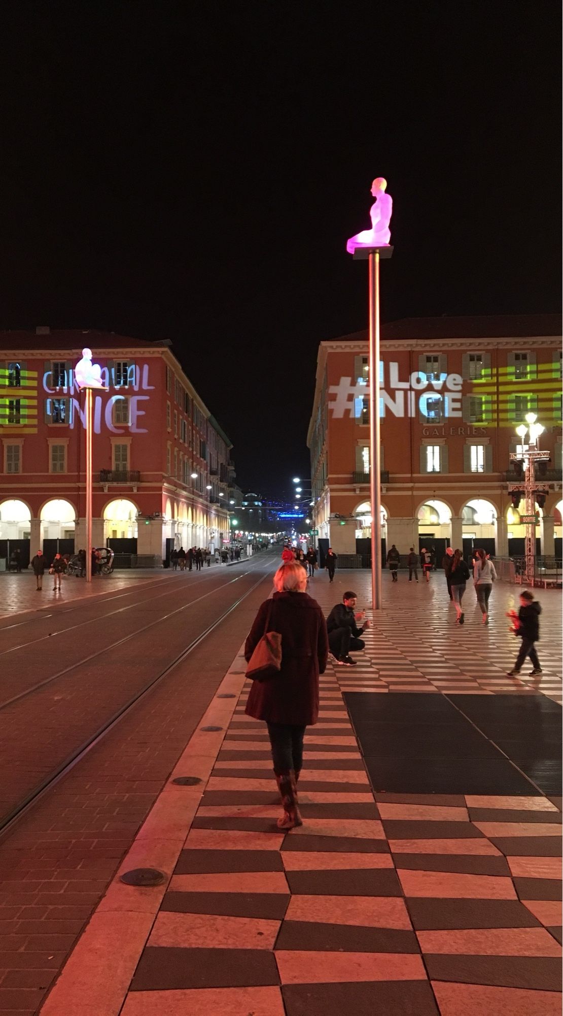 A Nice square with odd chequered paving and illuminated figures on poles.  My wife is walking away towards the far side of the square and the illuminated arched colonnades.  Projected onto the buildings: I love Nice.  Fair enough.