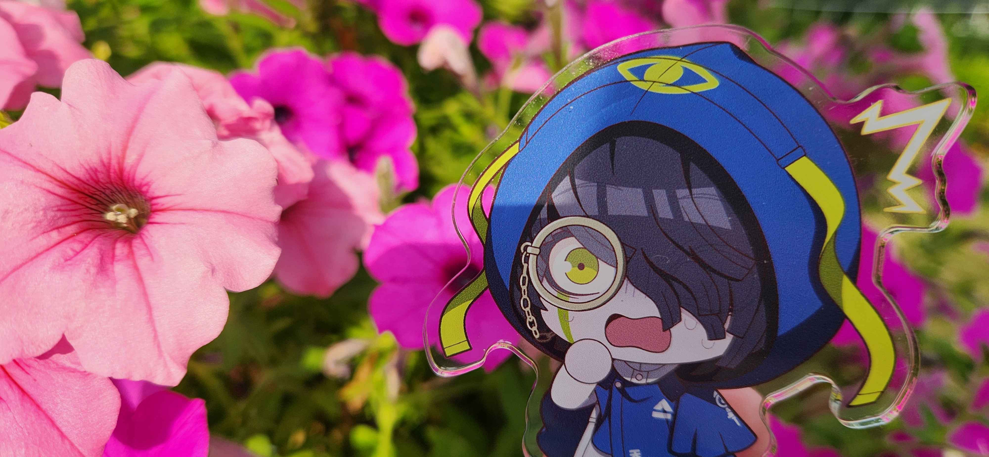 A photo of a chibi acrylic standee of the character Seth Burroughs. He's placed against some pink petunias, and it looks like he's startled by how large they are.