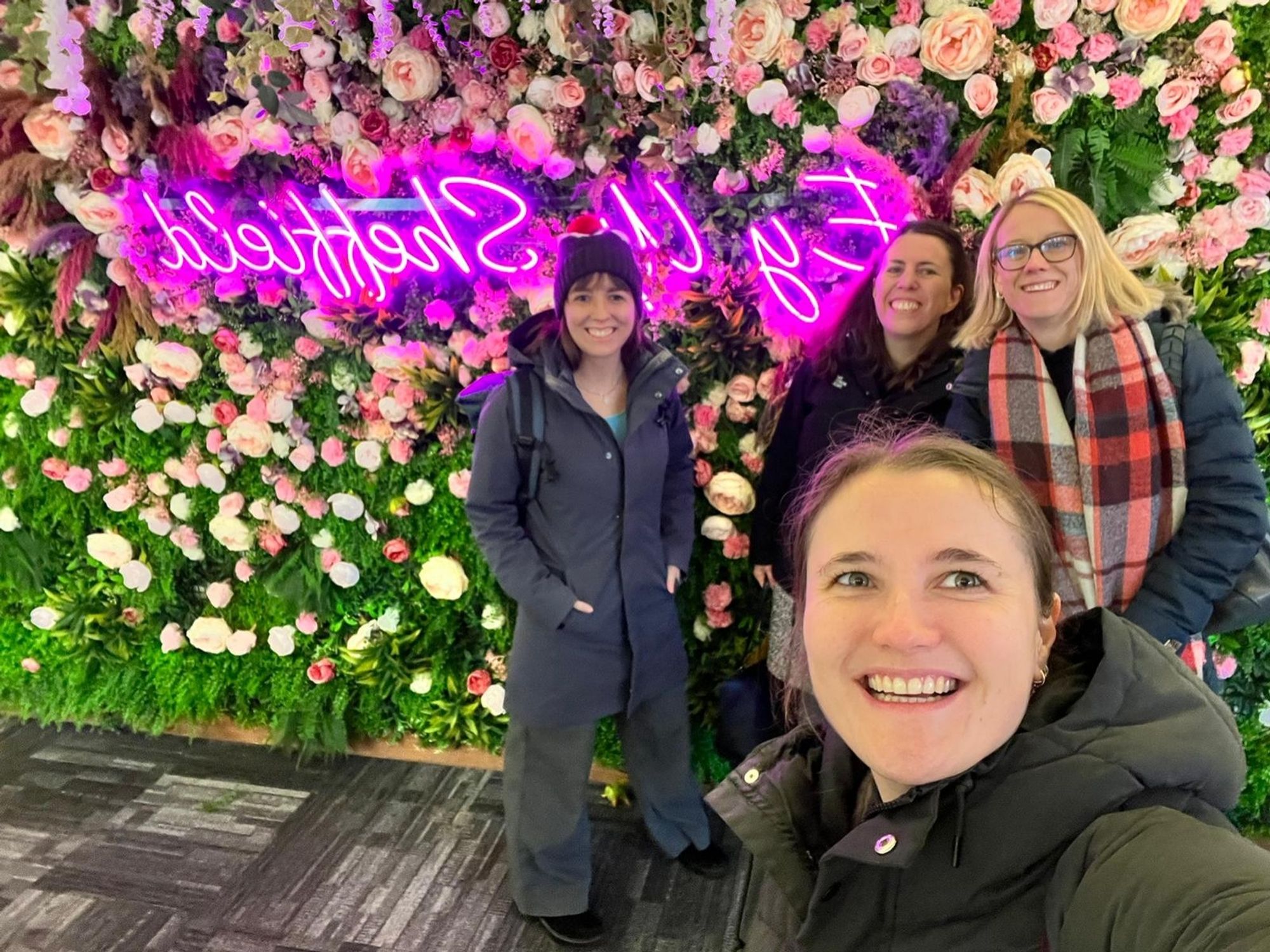 Caro Hautekiet, Vanessa Loaiza, Claudia von Bastian, and Evie Vergauwe in front of a neon sign that says "Ey Up, Sheffield" which is local language for a friendly hello or watch out