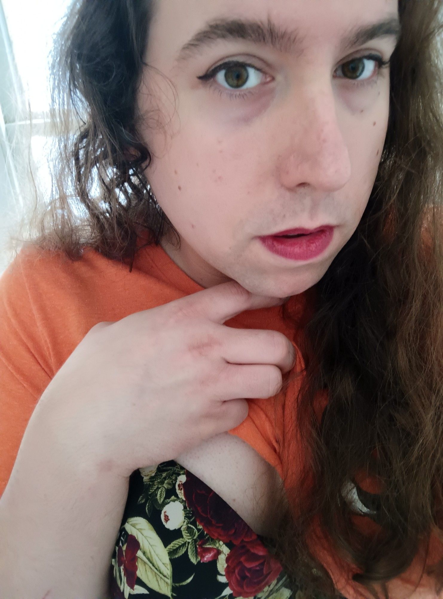 A woman with long brown hair slightly lifts her bright orange shirt, showing a floral bra underneath. She looks at the camera longingly, capturing a moment of tranquility and self-assuredness.