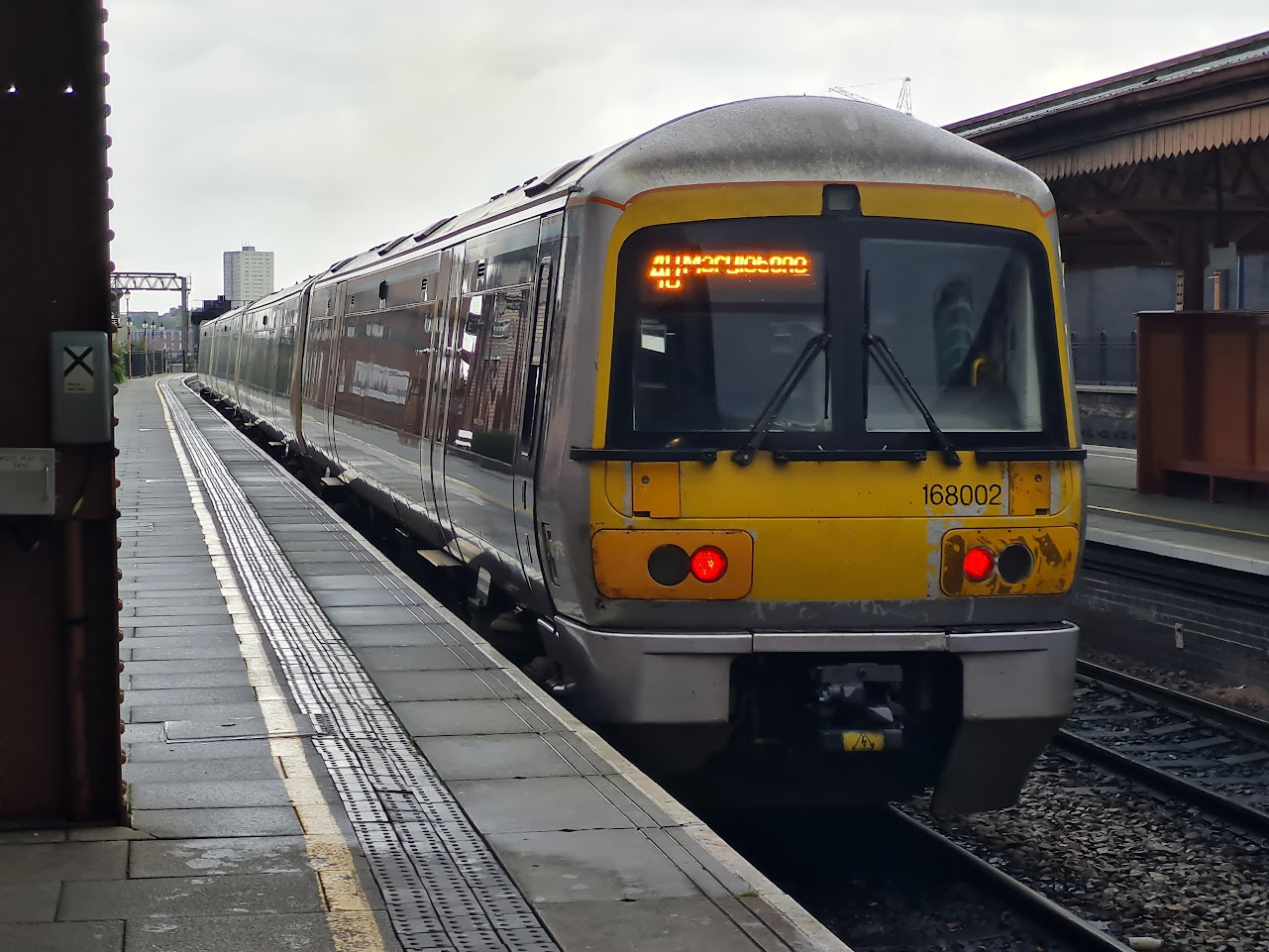 A photo of an Adtranz Class 168 'Clubman' Train in a Gray and Yellow Chiltern Railways livery, seen departing from a platform at Birmingham Moor Street railway station. The platform is damp, and the front of the train has some levels of wear and tear. The sky is gray.