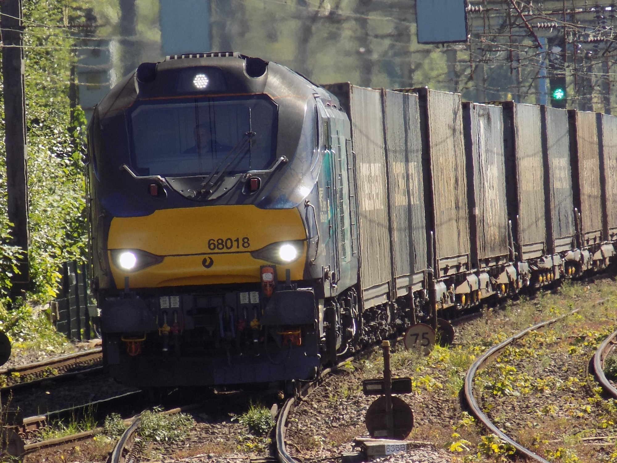 A zoomed-in Stadler UKLight Class 66 freight train in a Direct Rail Services livery, with a bright yellow front, seen towing numerous wagons of freight containers. The train is seen approaching Preston station heading northbound towards Scotland.