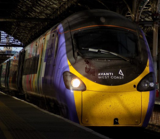 A zoomed-in, low contrast photo of an Alstom Class 390 Pendolino train, in Avanti West Coasts 'Progress' livery. The train is seen under the iconic roof at Preston Railway Station, as it boards passengers heading from Scotland to London.