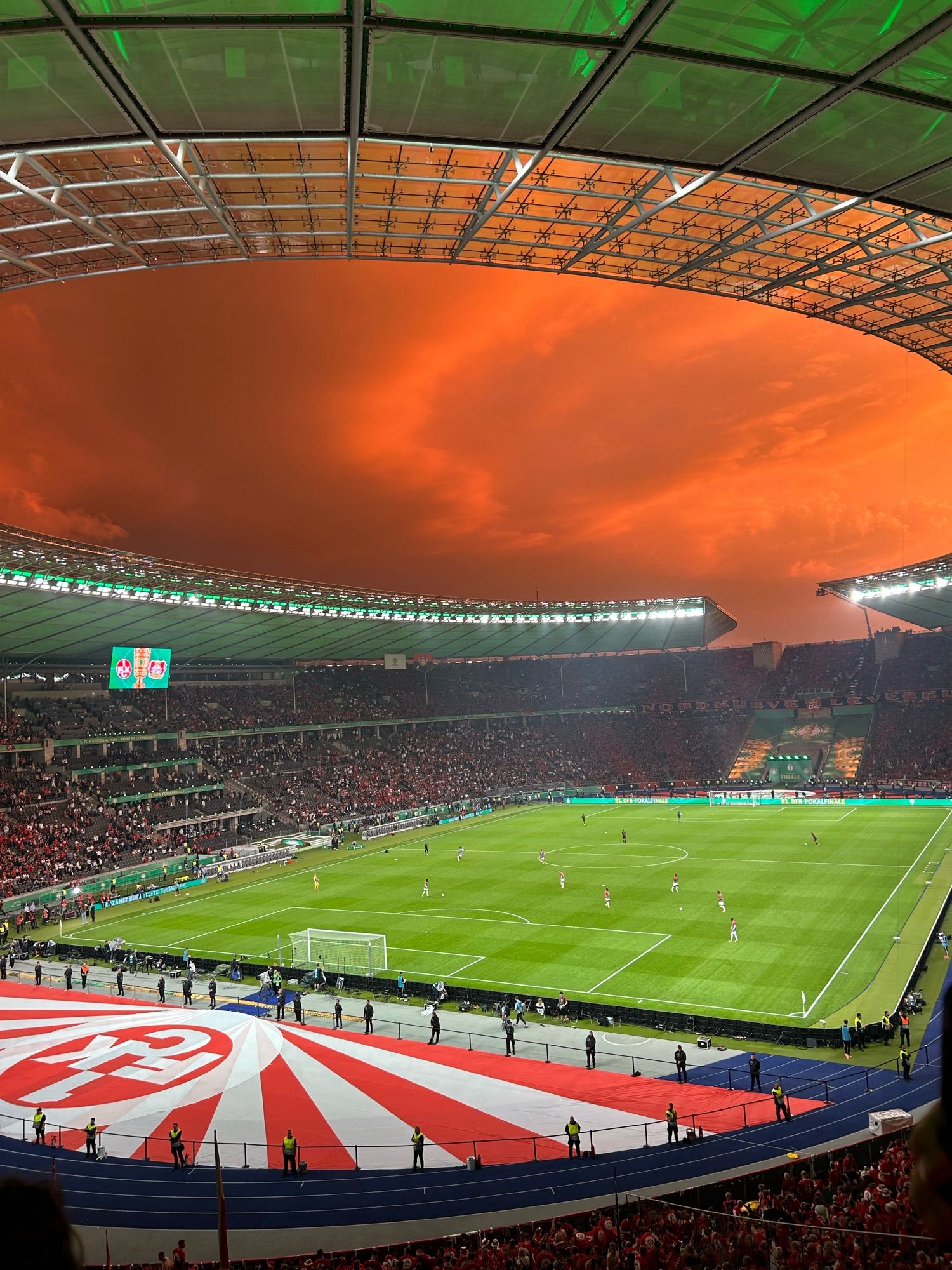 Ein Blick im Olymiastadion auf den Rasen, rot-weiße Banner am Boden, der Himmel ist bewölkt, die Wolken leuchten orange