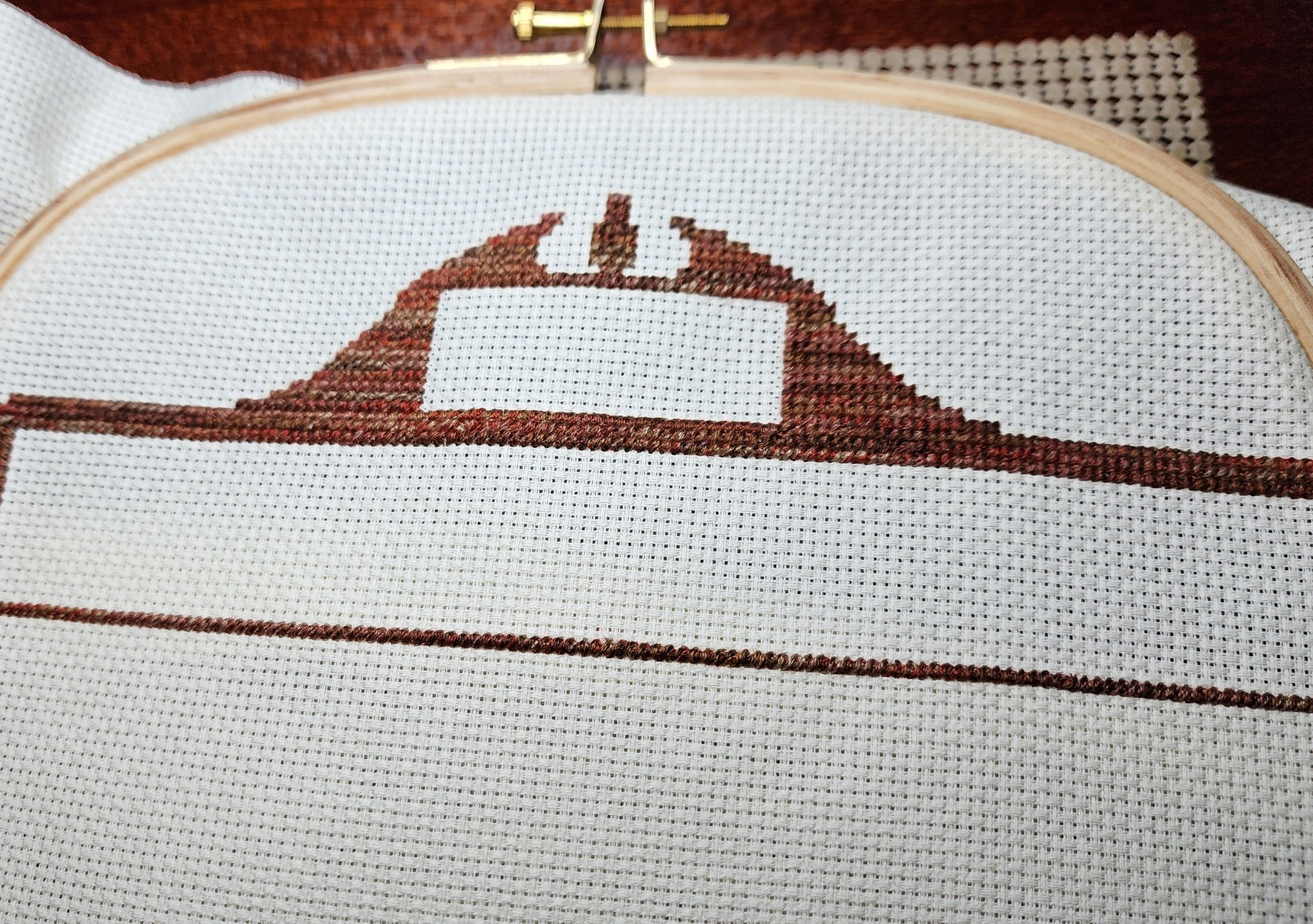 A square embroidery hoop is shown holding a canvas. On the canvas is the start of a bookshelf, cross stitched with variable floss in shades of brown. 