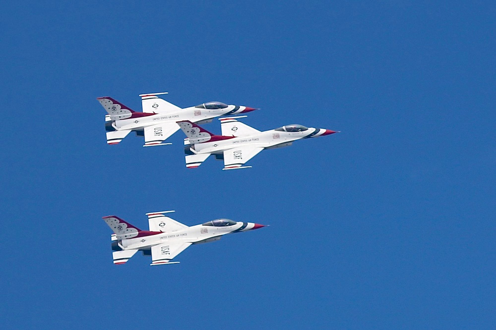 Three mostly white fighter planes with red and blue accents painting fly in formation from left to right against a deep blue sky.   Precision, danger and technology on display.