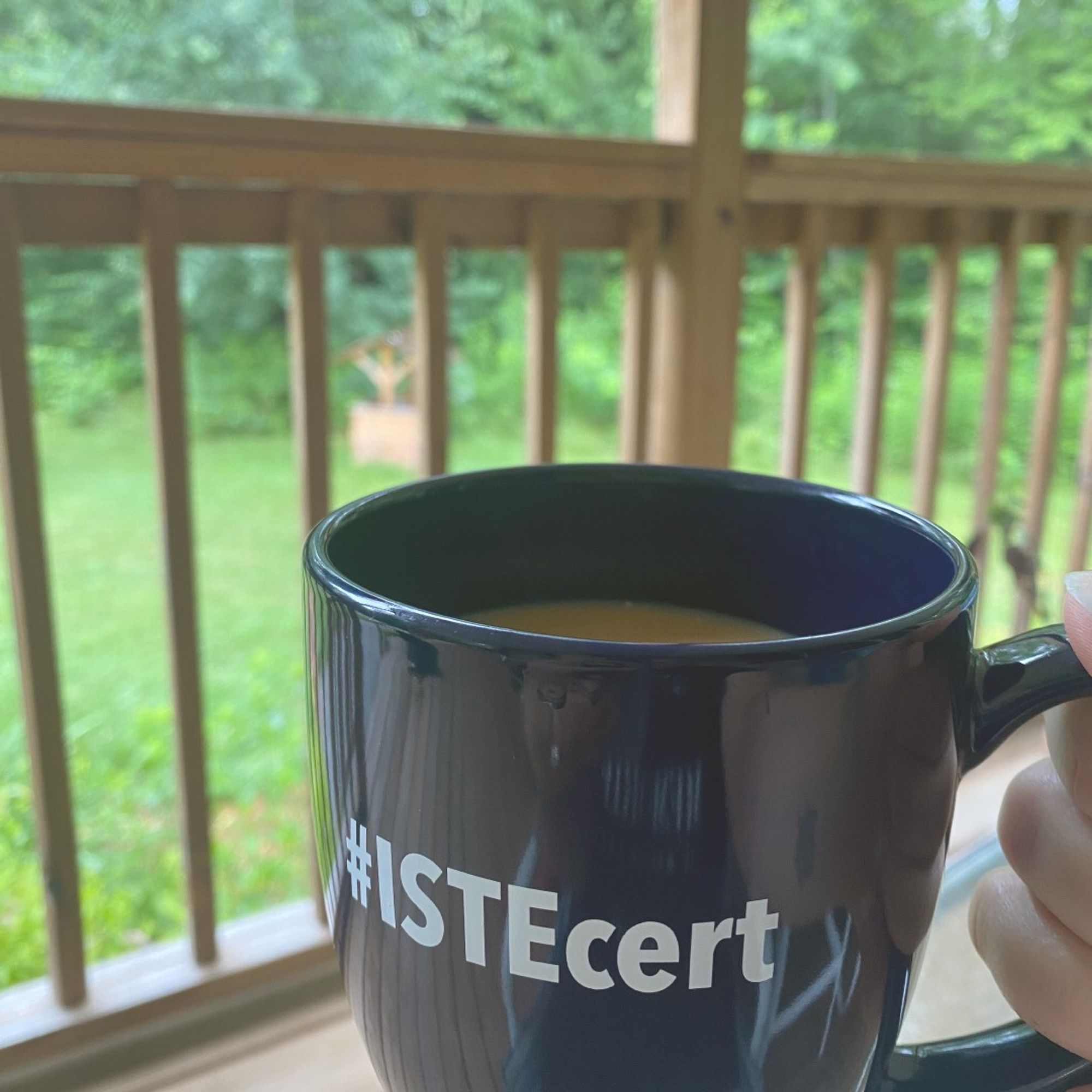 natural wood front porch, coffee cup with hashtag "ISTEcert" held up with green wooded area in background