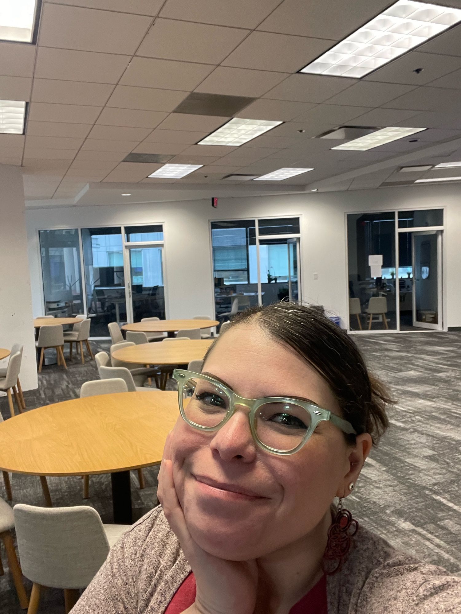 OP, smiling with chin resting on hand, in front of an empty room of tables with darkened offices in the background.