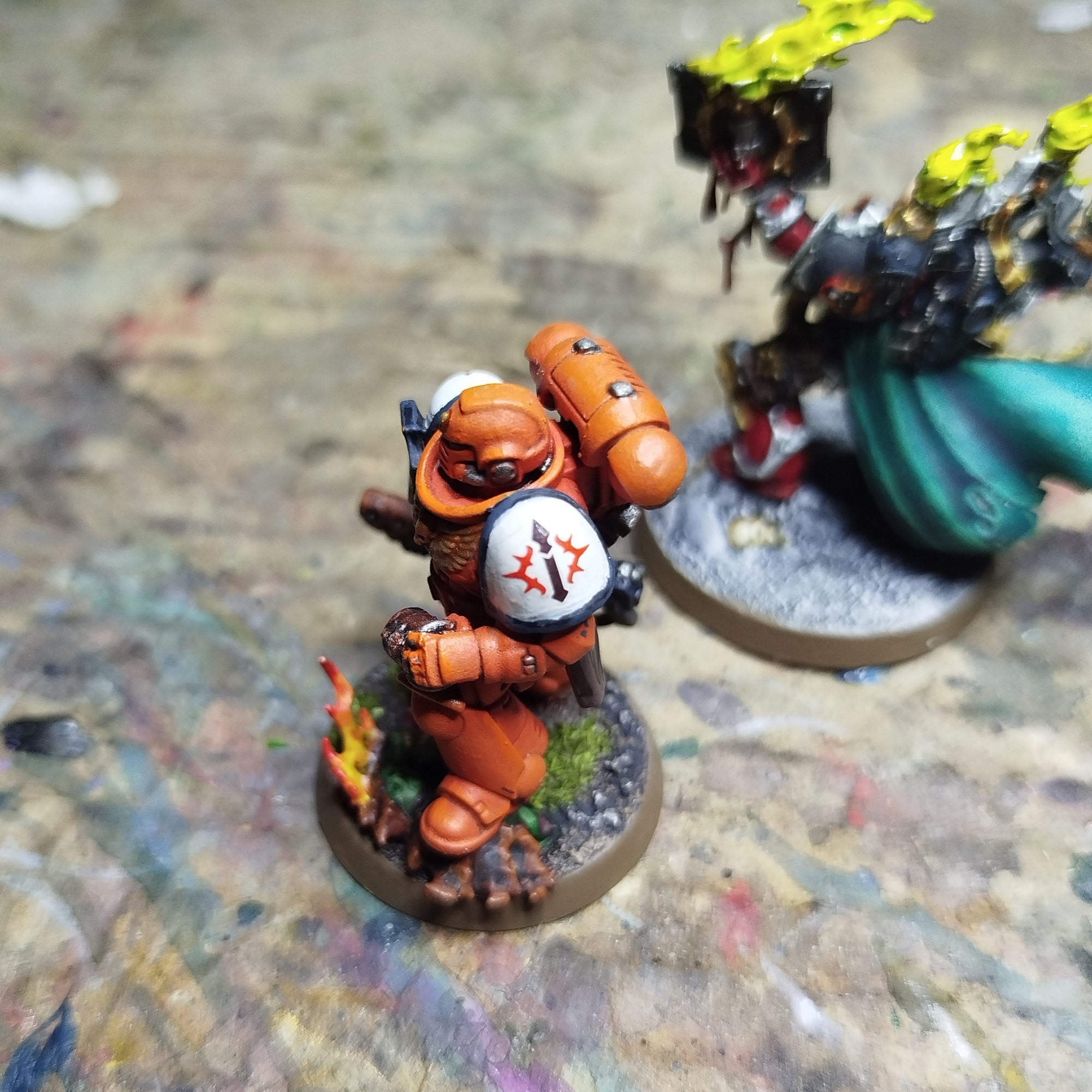 Foto de una miniatura de un marine espacial sobre el cartón que uso para pintar.

El marine esta pintado de color naranja con las hombreras de blanco adornadas por un reborde de color negro.

La foto hace énfasis en la hombrera para resaltar la calcomanía de una lanza negra rodeada por una aureola roja que hace de icono del capítulo para este marine.