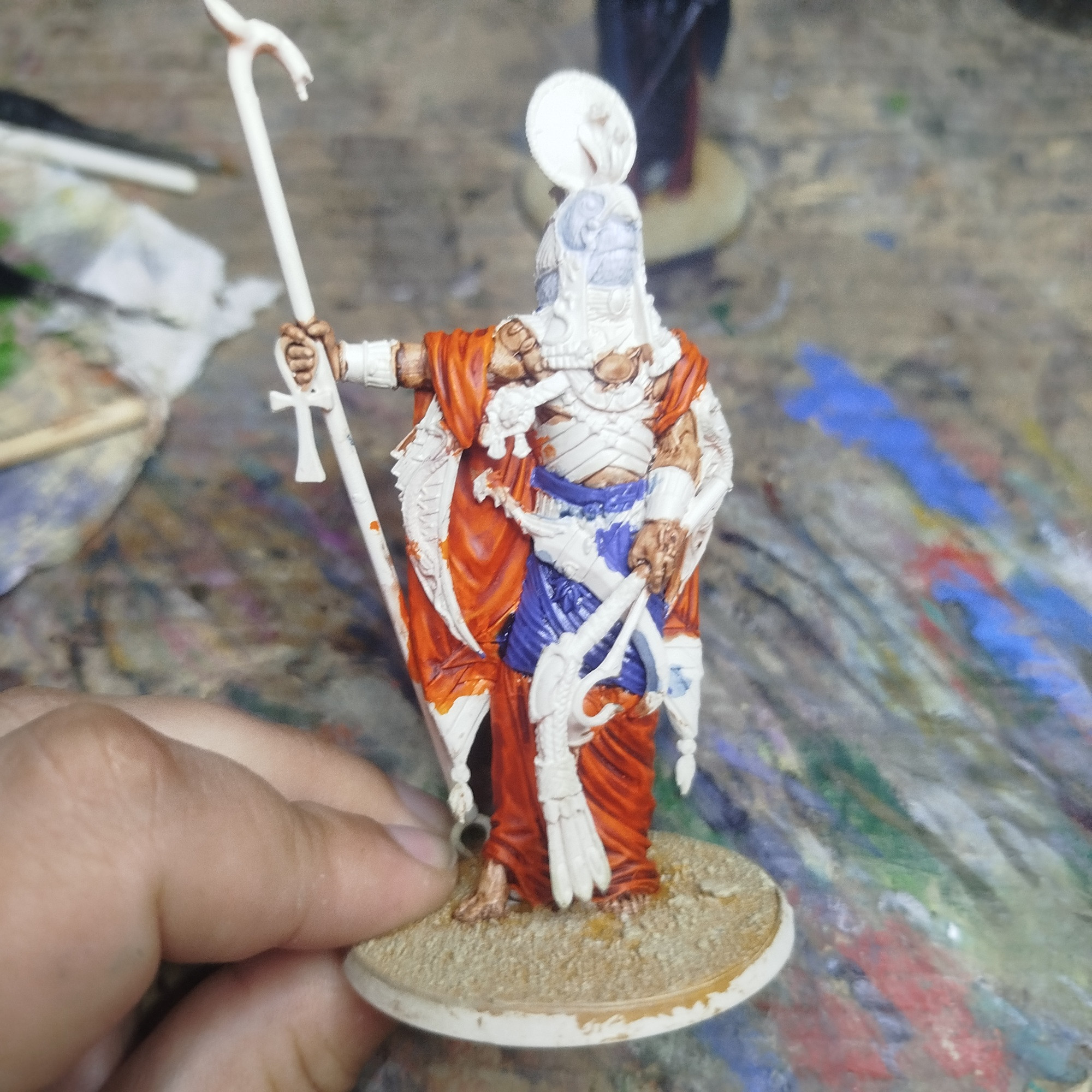 Foto de mi mano sosteniendo una miniatura de Ra para el juego de mesa Ankh a medio pintar.

Su túnica está pintada de naranja y azul y su peana está decorada con una temática de desierto.

El resto de la miniatura aún se ve de color blanco por la imprimación usada.