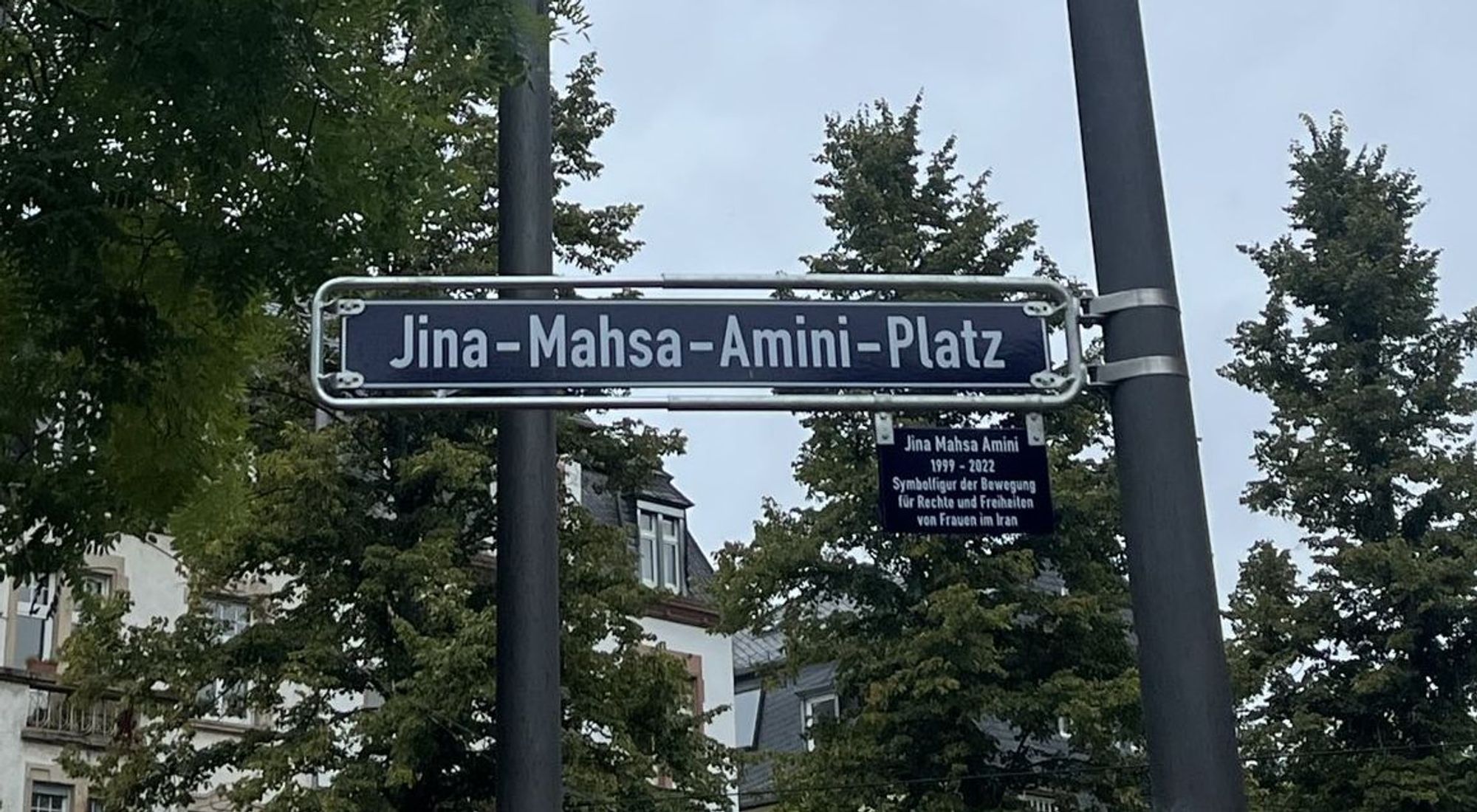 Foto des Straßenschildes Jina-Mahsa-Amini-Platz, darunter eine kleine Erklärtafel: "Jina Mahsa Amini 1999-2022. Symbolfigur der Bewegung für Rechte und Freiheiten der Frauen im Iran"