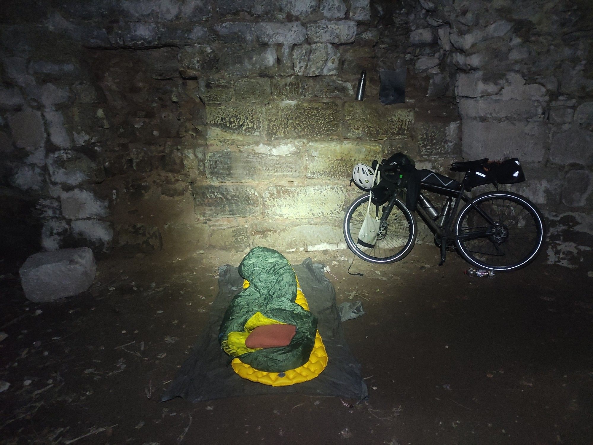 Blick auf eine dicke alte Steinmauer an der ein Fahrrad lehnt. Davor eine Isomatte und ein darauf ausgebreiteter Schlafsack.