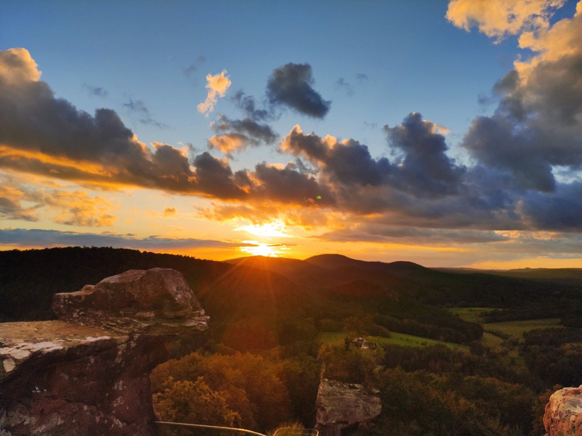 Sonnenuntergang hinter bewaldeten Hügel vom höchsten Punkt einer Burgruine die Wolken am Himmel sind golden eingefärbt von der Sonne.