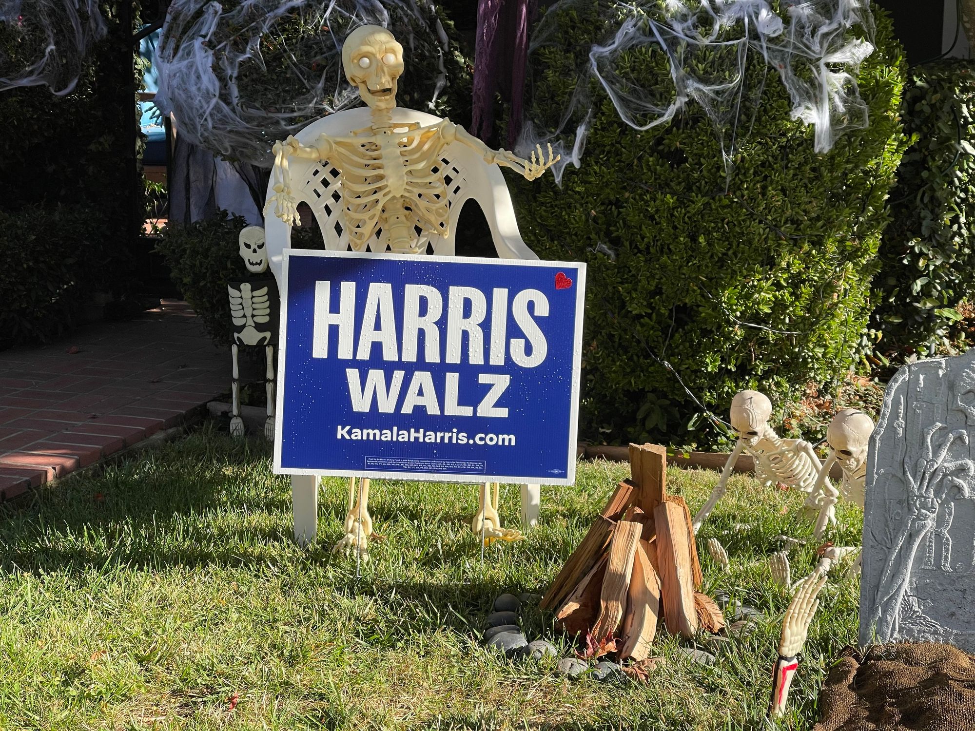 A halloween plastic skeleton sitting in a chair in front of a Harris/Waltz yard sign.