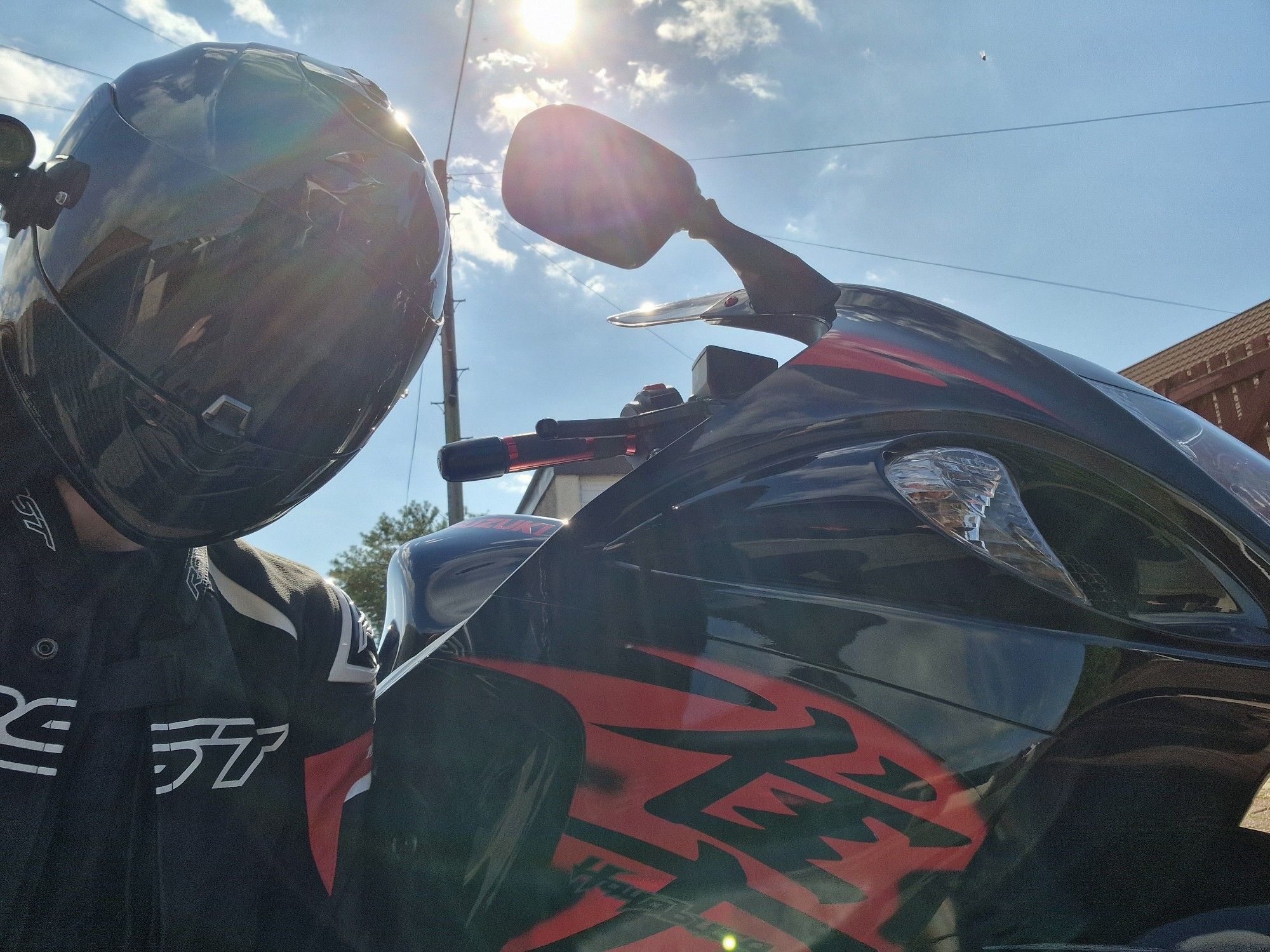 Selfie in front of a blue sky and bright sun, myself in a black motorcycle helmet crouched next to my red and black Hayabusa motorcycle.