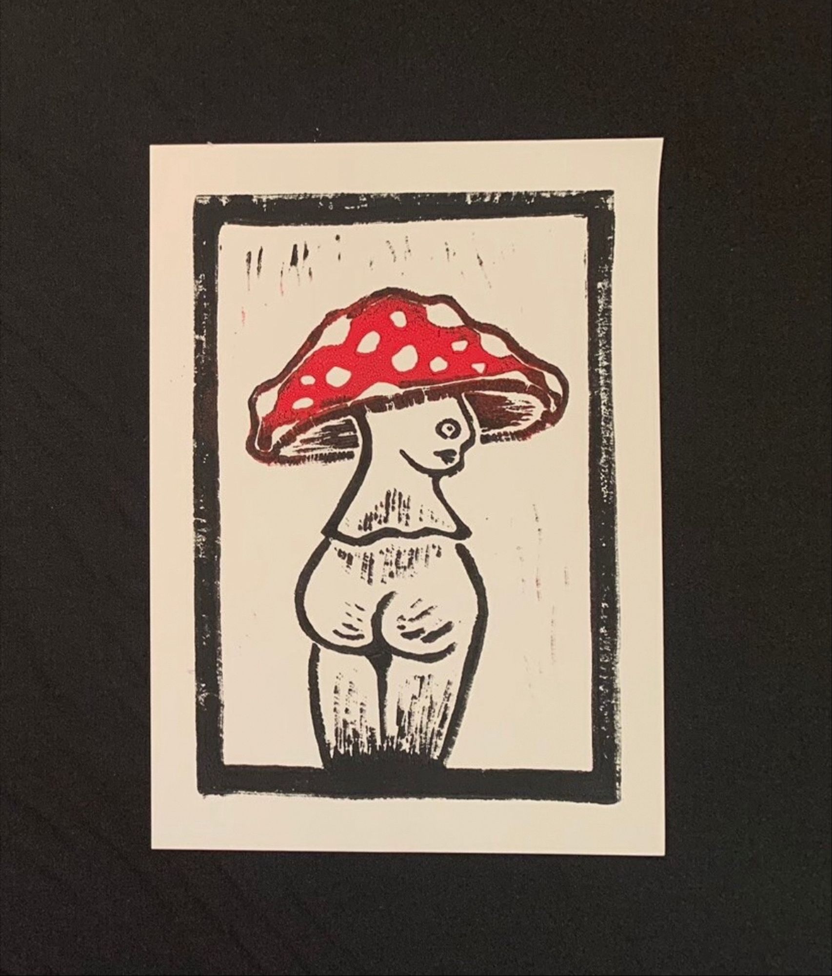 A photo of a hand printed linocut on white paper. Image shows a red mushroom with a face and large butt outline by a one centimeter black border.