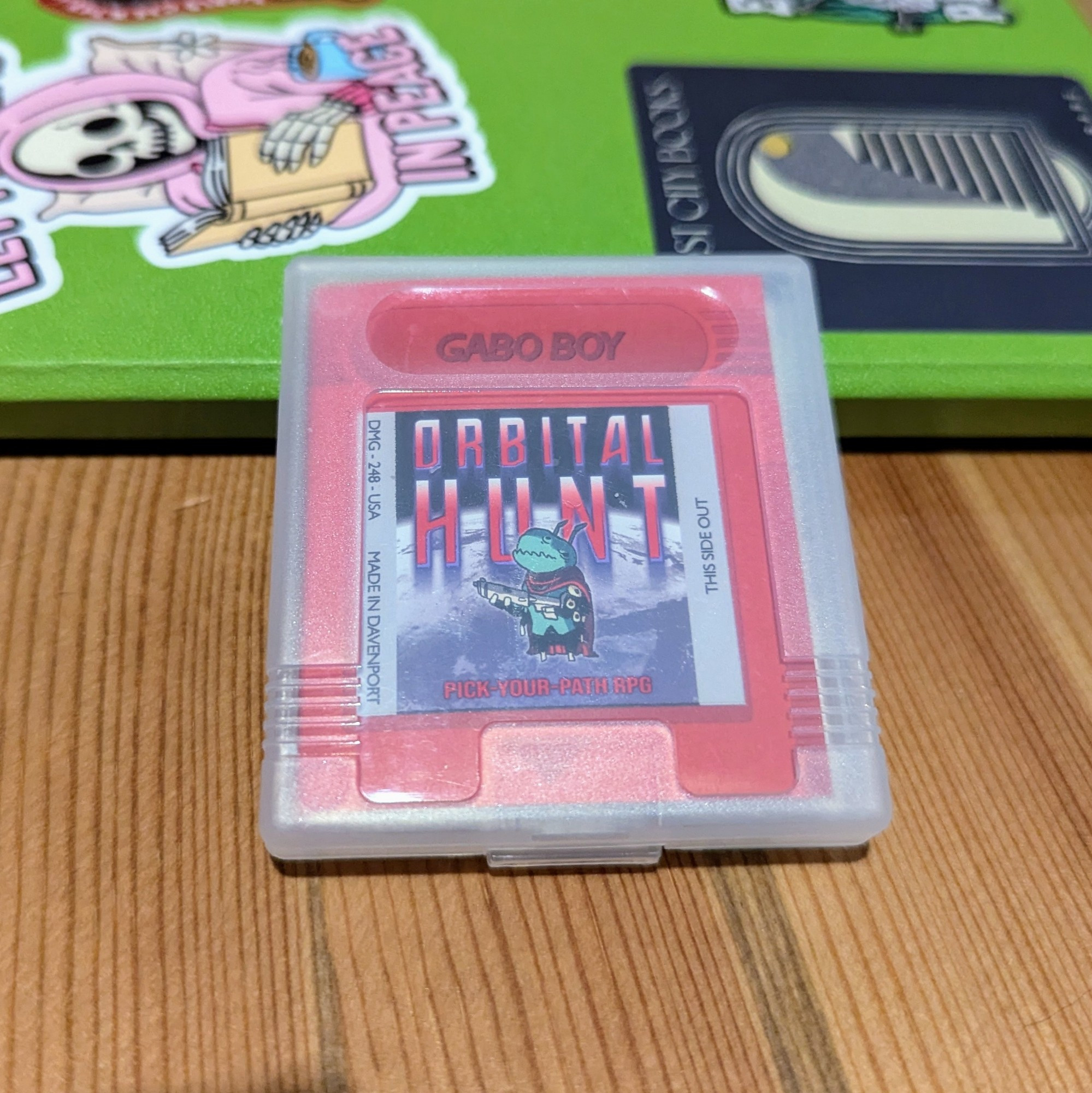 Photo of a tiny ttrpg zine called orbital hunt inside a game boy cartridge case. The zine is red is stylized like a game boy cartridge. There's an illustration of a green alien holding a gun on the cover