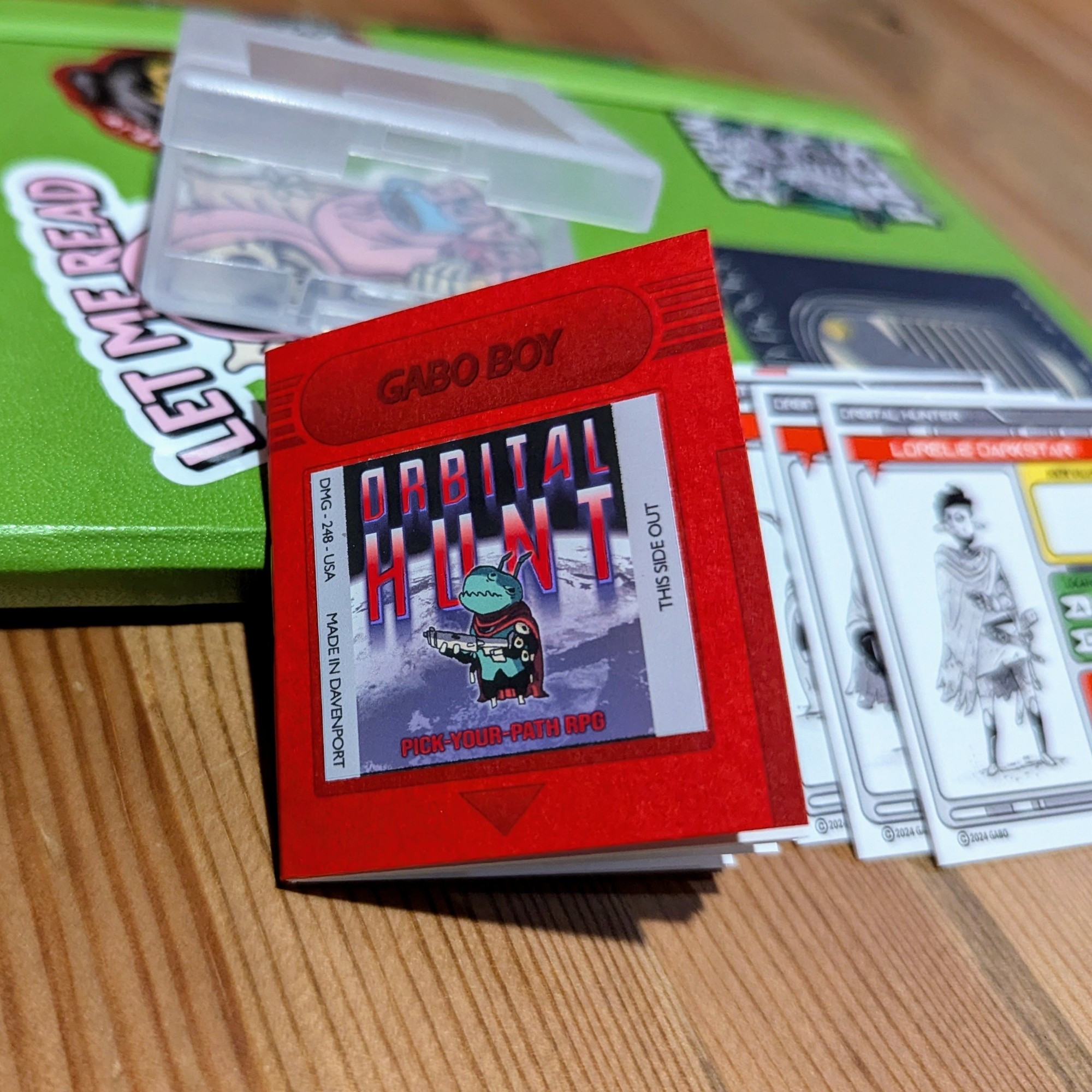 Photo of a small zine ttrpg called orbital hunt. It's stylized like a red game boy cartridge. There's an illustration of a green alien holding a gun on the cover.