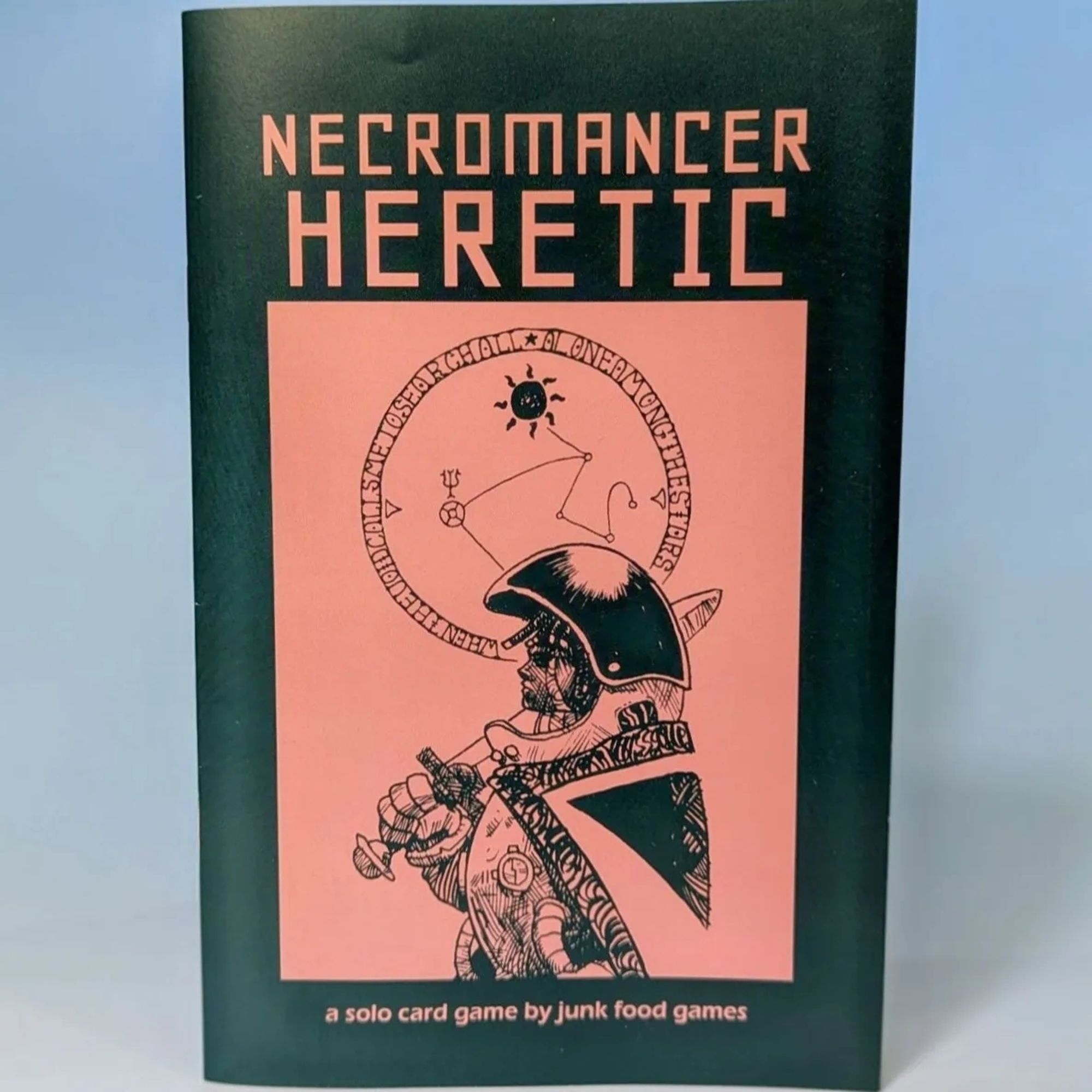 Photo of the game Necromancer Heretic. It shows the cover which has the title and an illustration of a sci-fi astronaut in profile holding a sword. There is some astrology symbols above them. At the bottom, it says "a solo card game by junk food games"