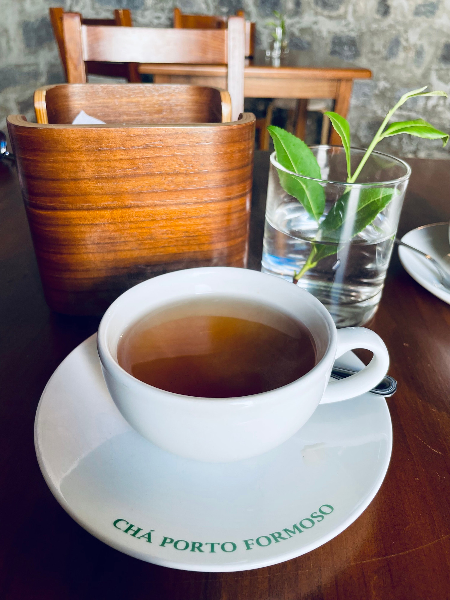 Cup of tea on table. Sprig of tea in glass as decoration. Stone wall in background. 