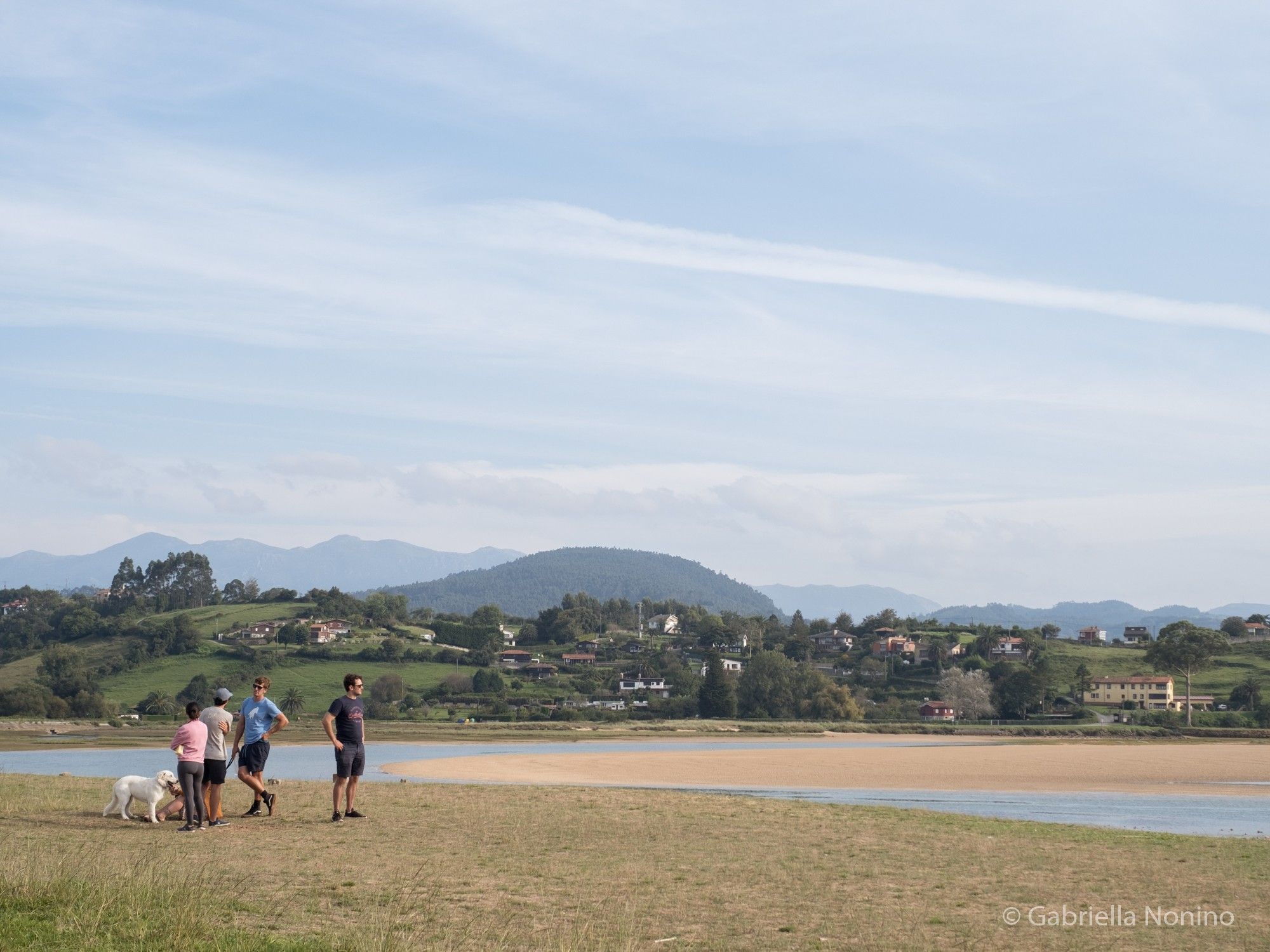 Four people and a dog on a sandy shore of the estuary of a river.