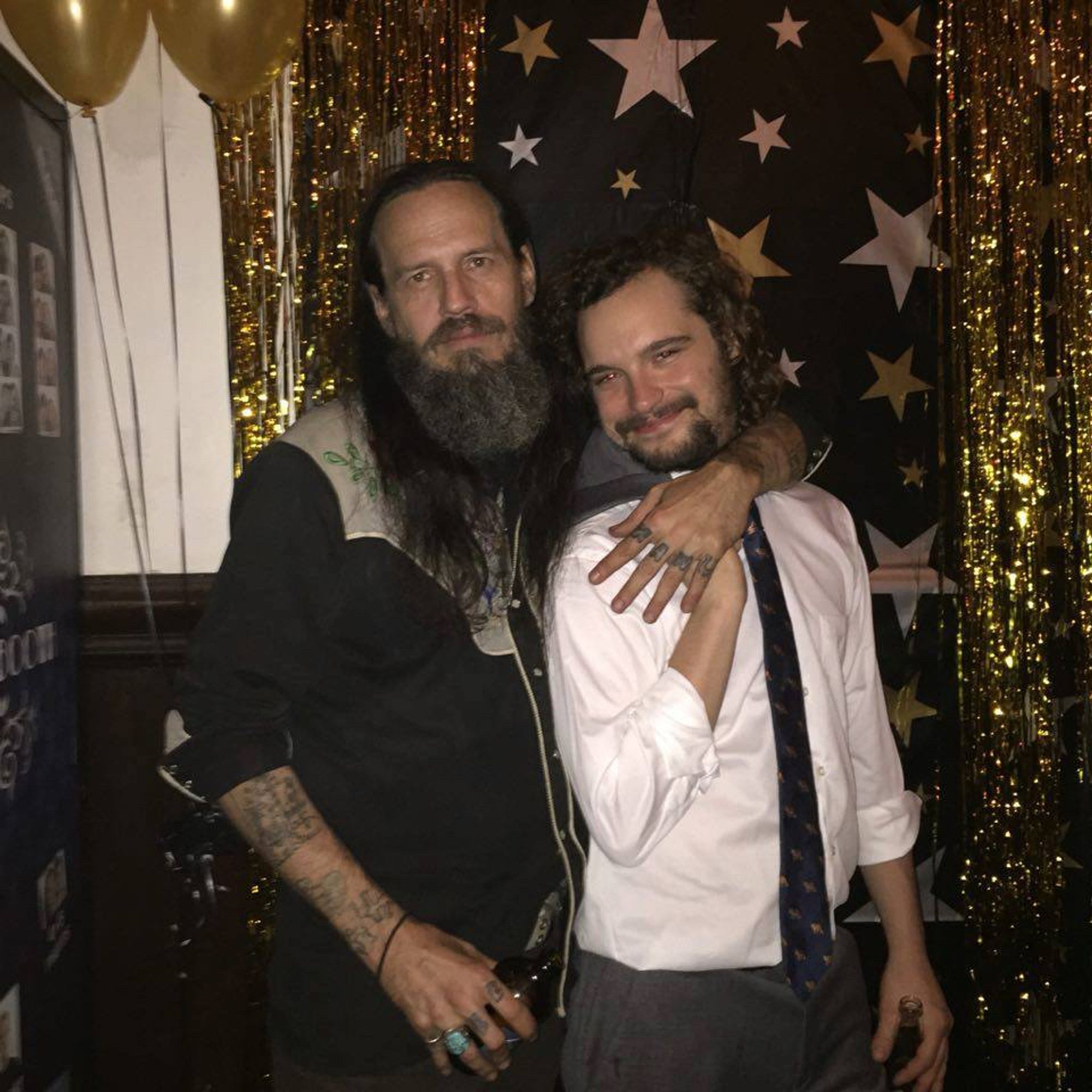 me in a vintage embroidered western shirt, i have long hair & beard & i've forgotten my glasses somewhere, i'm hugging a dear friend of mine who is wearing a suit & tie. we're both holding bottles of beer & are posing in front of a gold and black shiny background