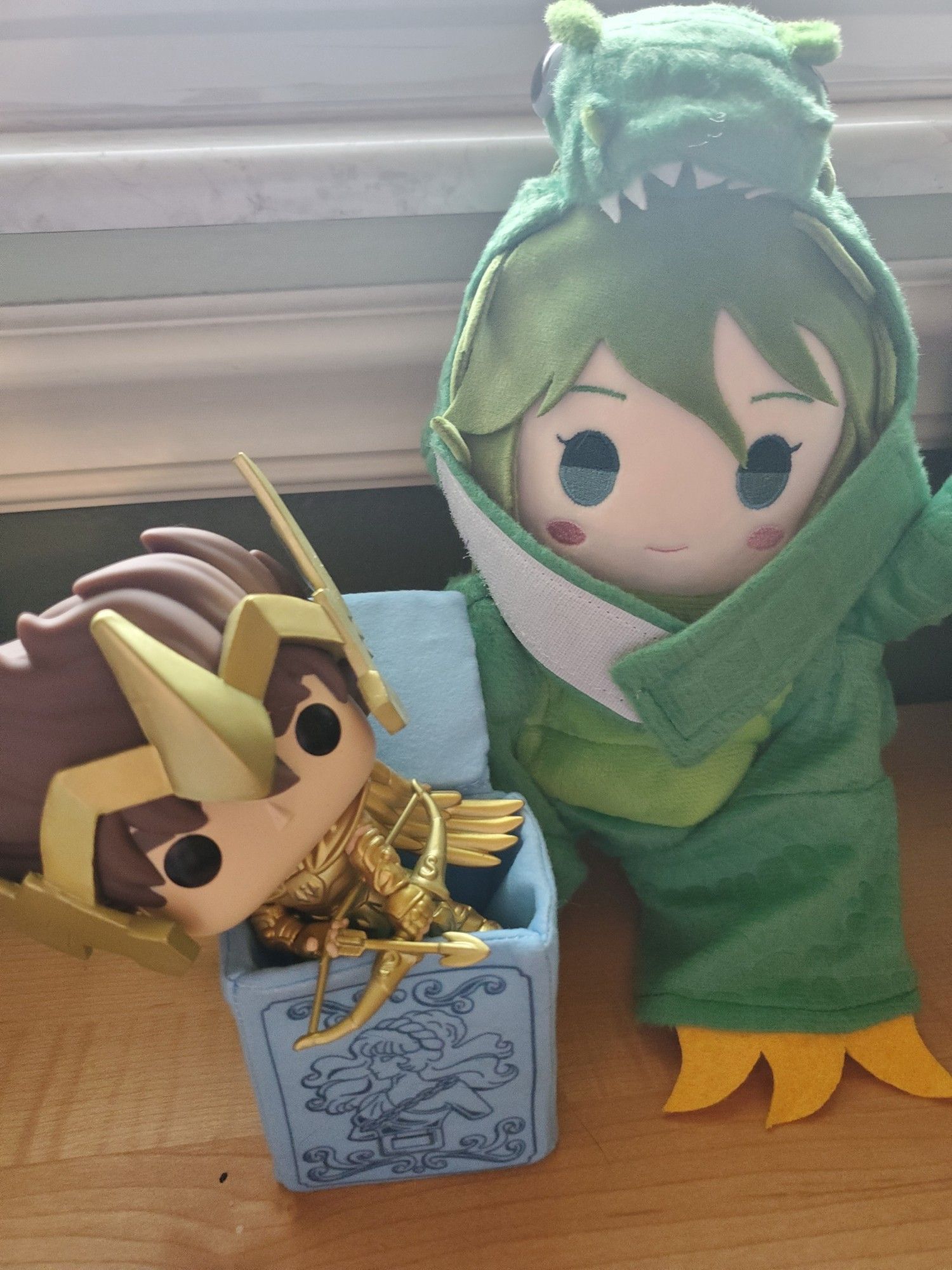 Andromeda Shun plushie wearing a dino costume.  In his Pandora box, the Sagittarius Seiya funko pop is posing