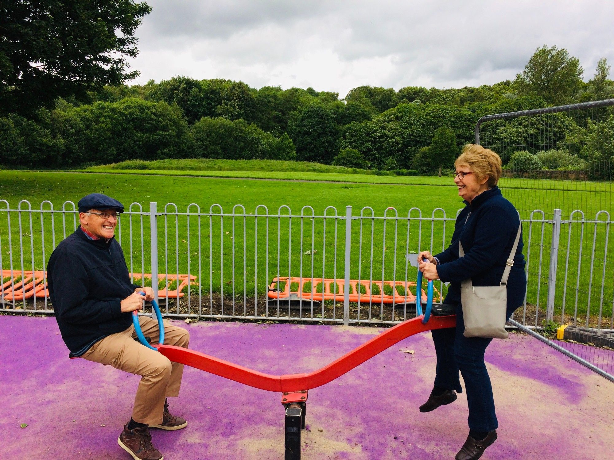 Two adults on a playground see saw.  The man low down and smiling at the camera.  The women is up and his smiling at him.