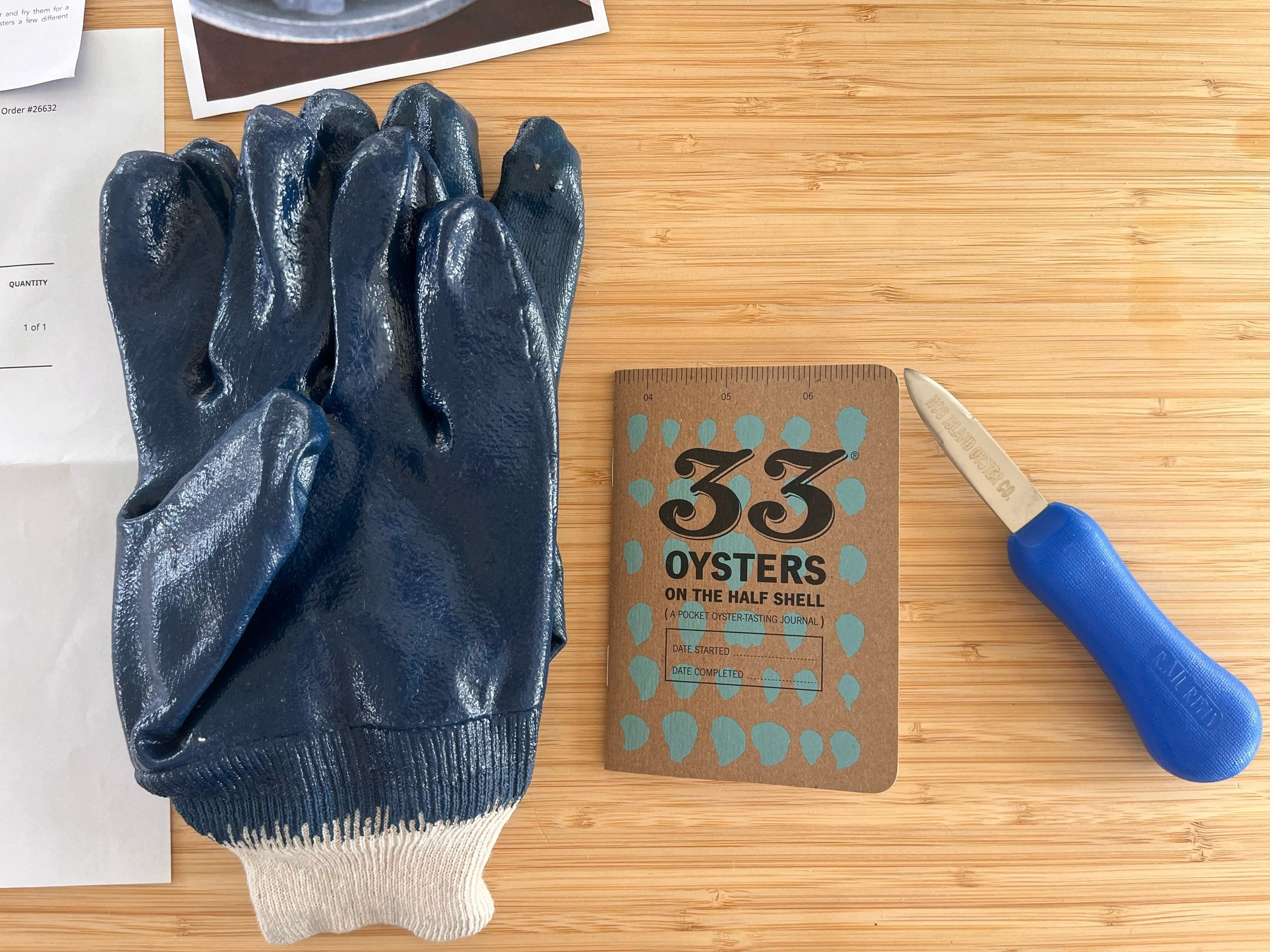 A picture showing a pair of shucking gloves, a “33 oysters on the half shell” tasting booklet and a shucking knife with the “Hog Island Oyster Co.” on the blade. All objects are sitting on a bamboo kitchen island.