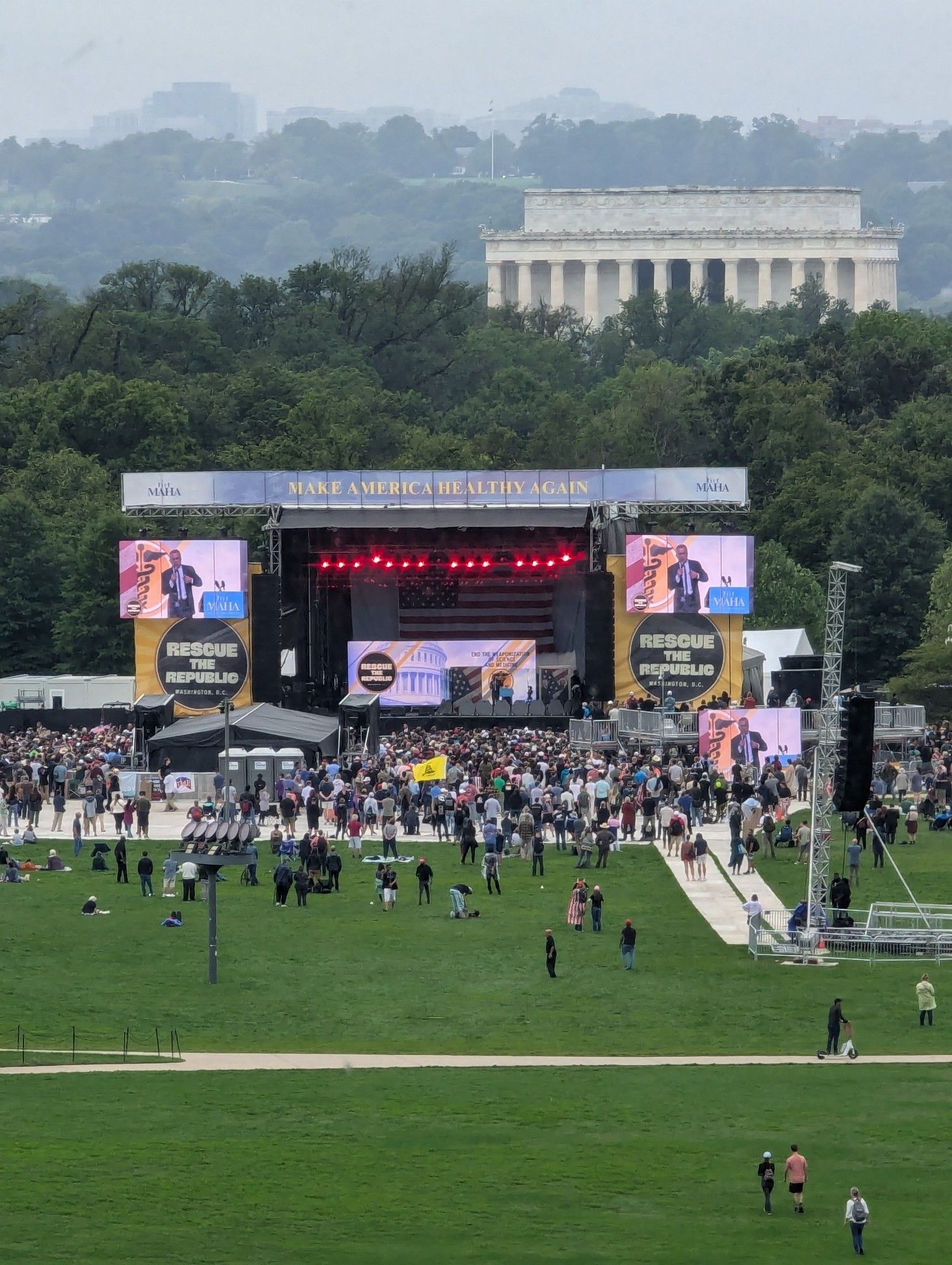 A small group of people in front of a large stage at the national mall with some trees and the Lincoln memorial in the background. Signs on the stage say "Rescue the Republic" and "Make America Healthy Again"