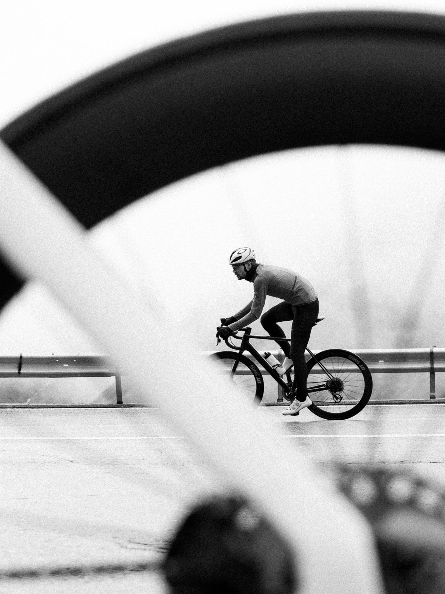 Schwarz-Weiß-Foto eines Rennradfahrers, der durch das Rad eines anderen Fahrrads hindurch fotografiert wurde. Das Rad im Vordergrund sieht man nur angeschnitten und verschwommen.