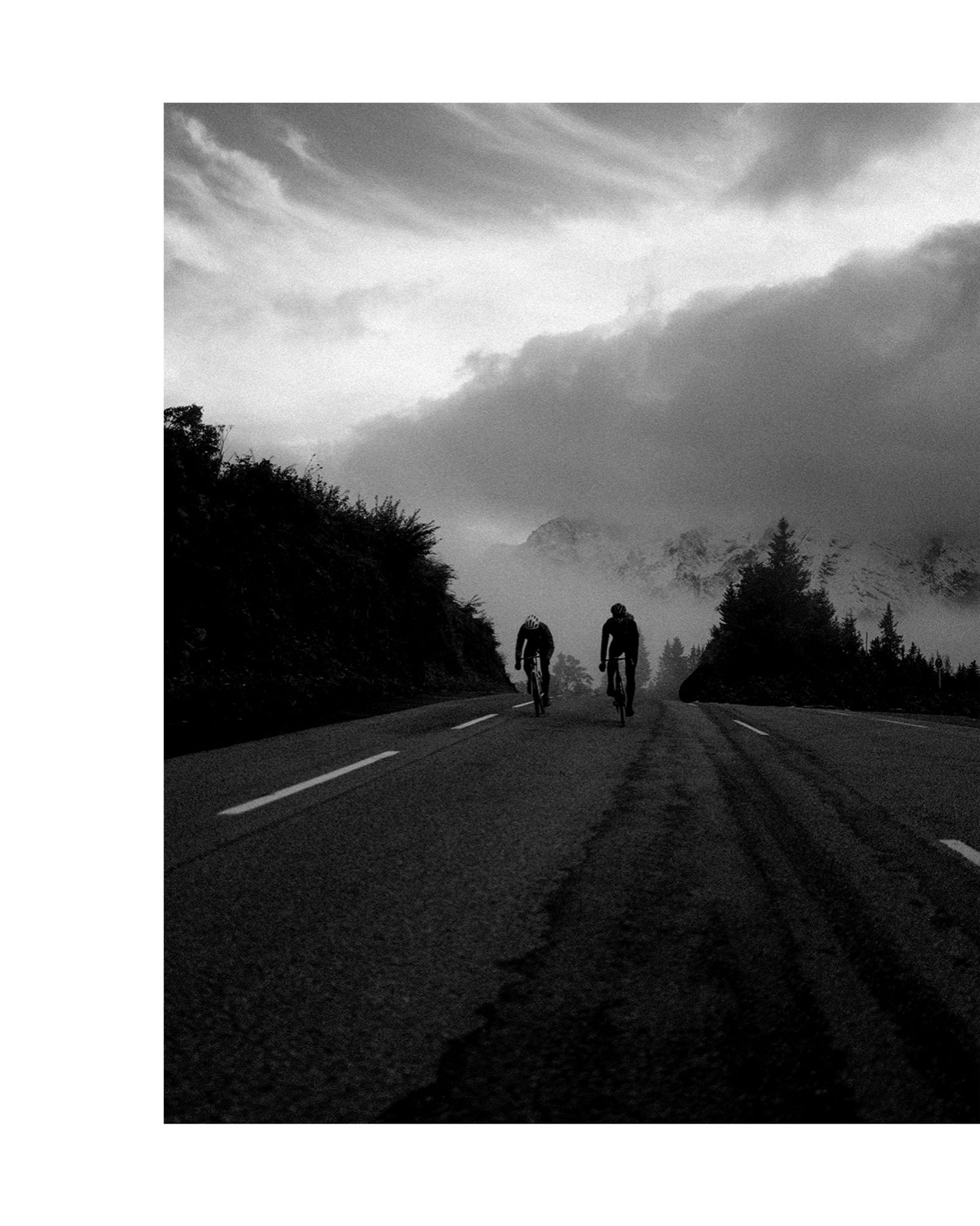 Schwarz-Weiß-Foto von zwei Rennradfahrern, die mittig im Bild auf einer asphaltierten Straße zu dem Betrachter entgegen fahren und in dem Moment einen Anstieg bewältigen. Im Hibtergrund sind mächtige schneebedeckte Berge zu sehen sowie links und rechts von den Radlern dunkle Nadelbäume.