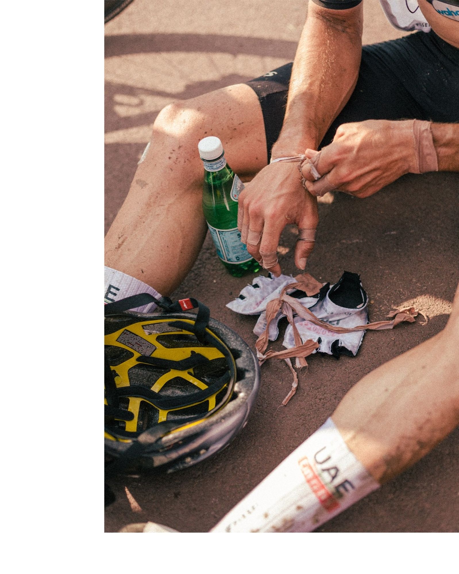 Ein Rennradfahrer sitzt nach dem Rennen Paris Roubaix auf dem Asphalt, man sieht nur seine Arme und Beine. Auf dem Boden zwischen seinen Beinen liegt sein Helm, seine Fahrradhandschuhe, eine Wasserflasche und das Tape, was er sich gerade von seinem rechten Handgelenk abwickelt.