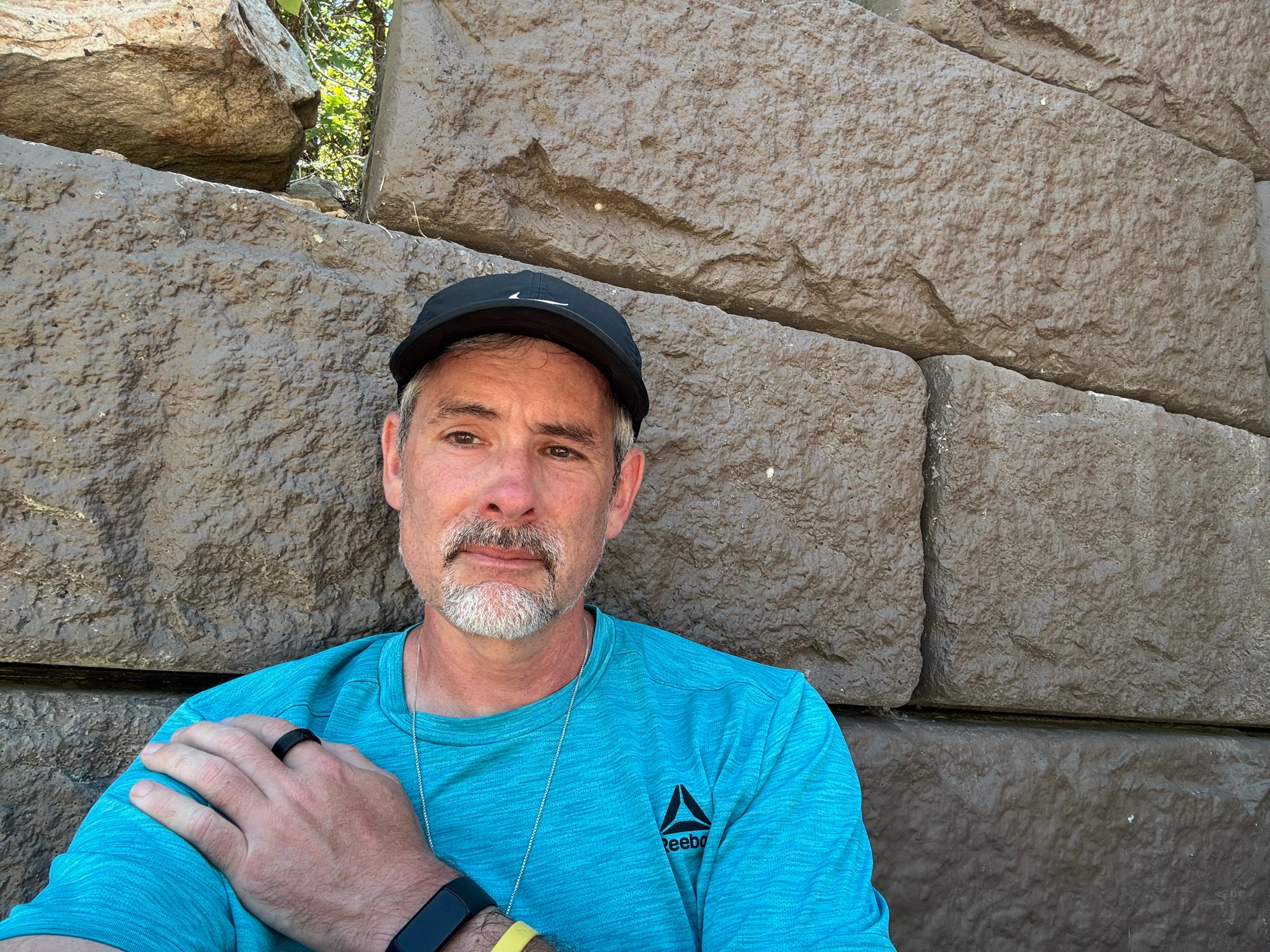 An eel historian in a blue shirt and black hat leaning against a stone wall. His left hand is in his right shoulder, and he looks thoughtful. 