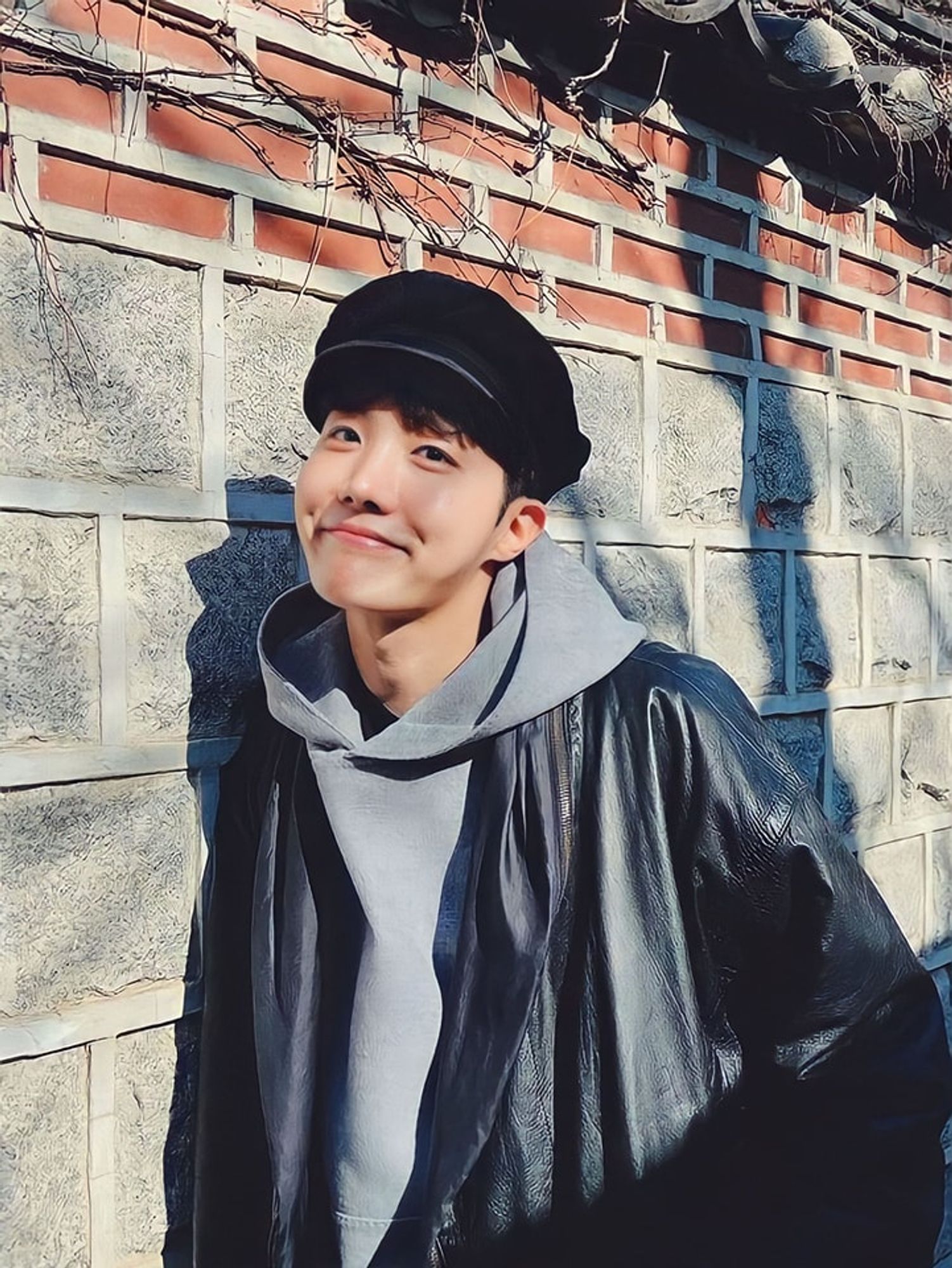 J-Hope leaning again a brick wall, leather jacket layered over a grey hoodie. He is wearing a black cabbie cap and a cute smile with bright eyes.